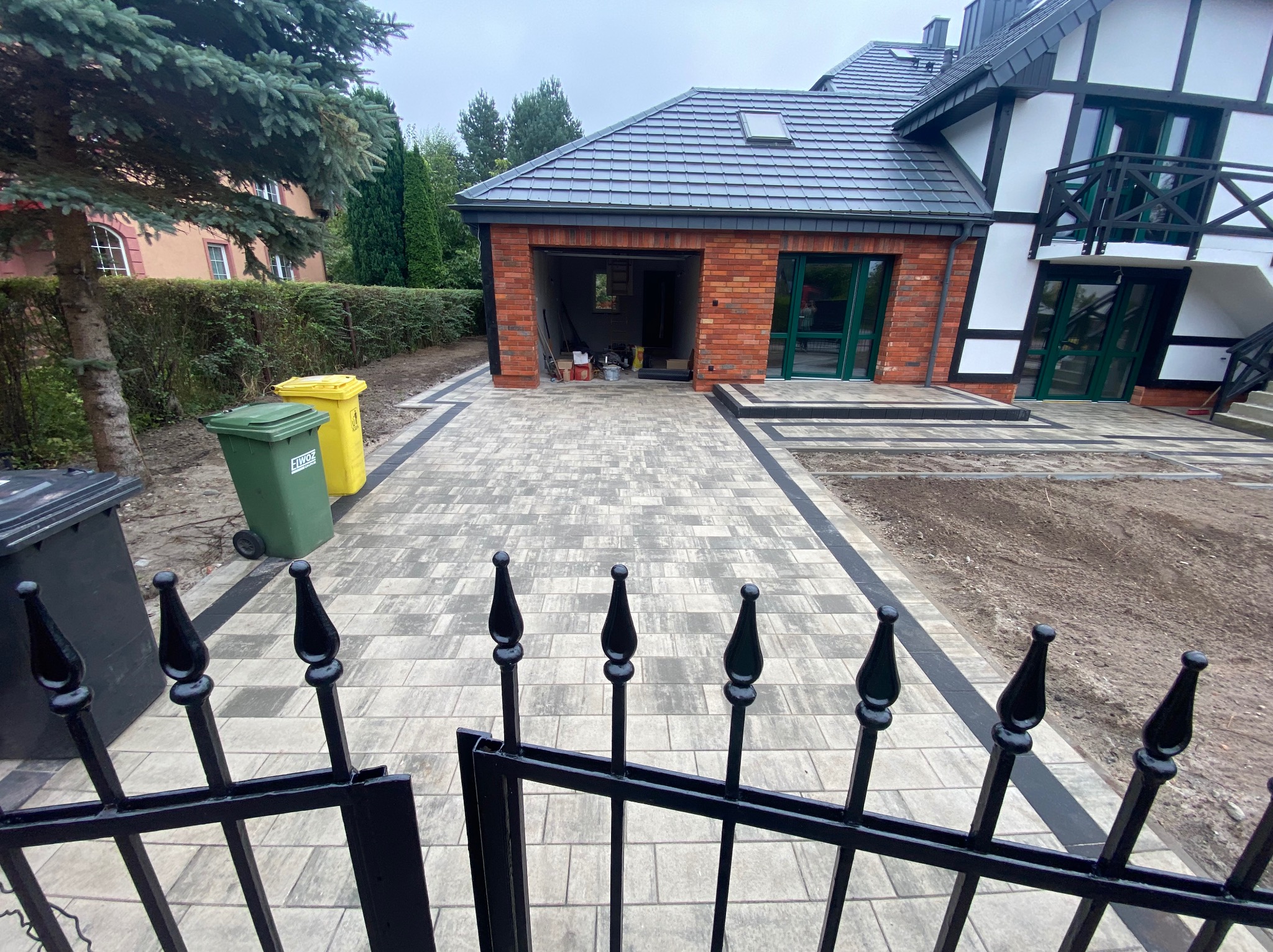 Ułożona kostka brukowa w odcieniach szarości z czarnymi obrzeżami prowadząca do garażu z cegły i domu z balkonem, widok zza czarnego, metalowego ogrodzenia.