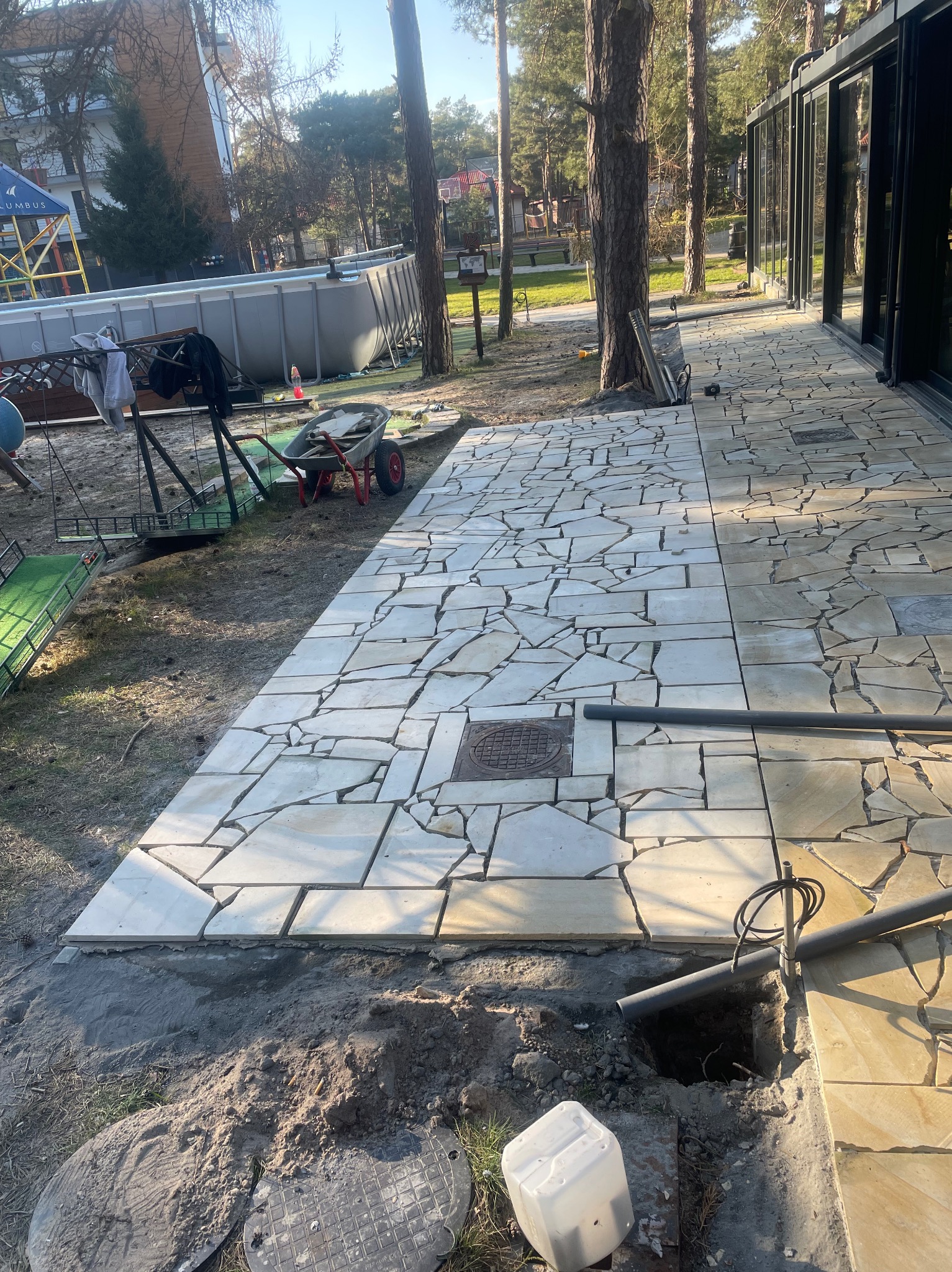 Fragment nowo ułożonego chodnika z nieregularnych płyt kamiennych w odcieniach beżu, z widoczną studzienką kanalizacyjną i rurami w trakcie montażu obok tarasu z przesuwnymi szklanymi drzwiami,...