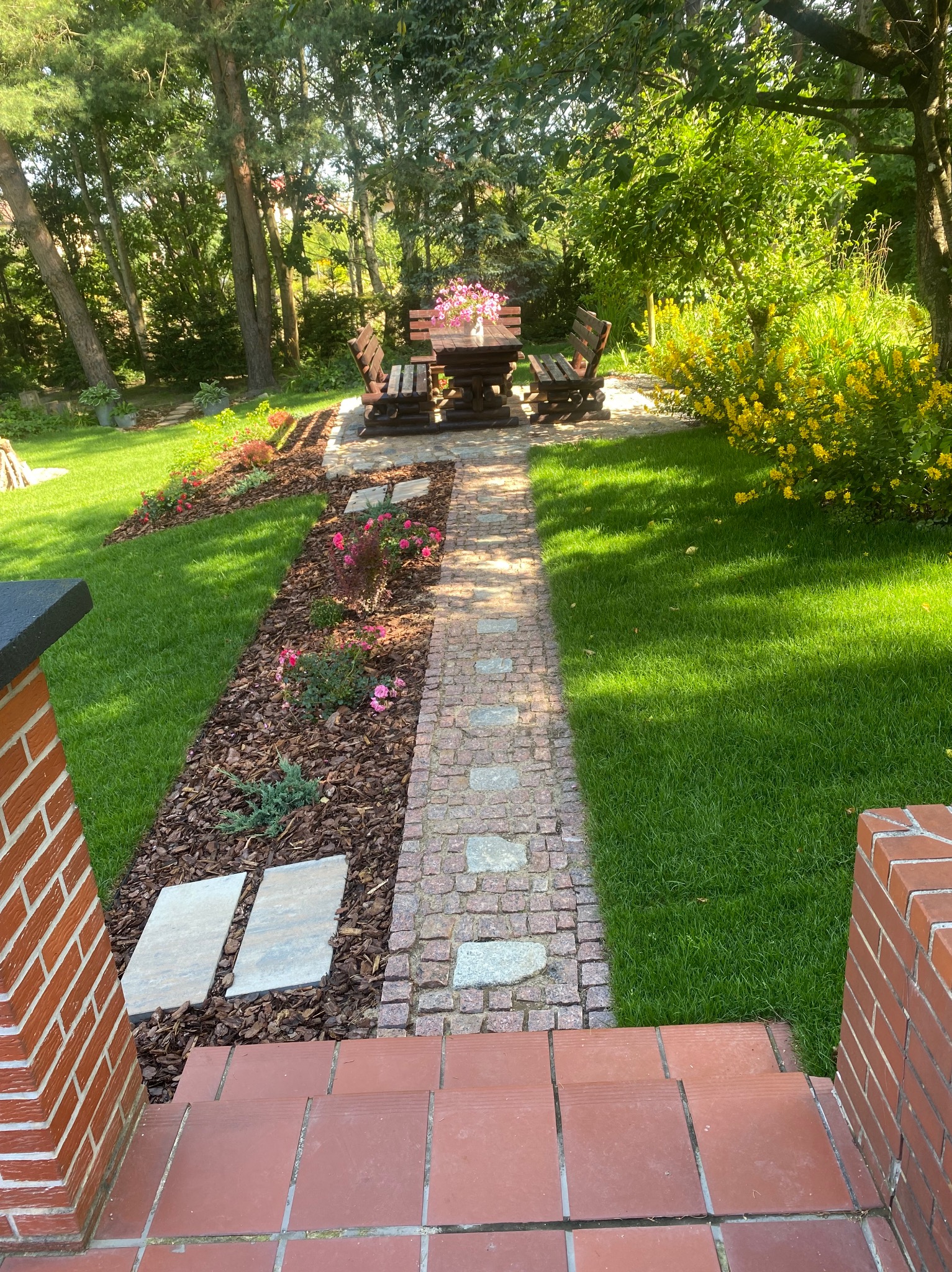 Ogród z ścieżką z kostki granitowej prowadzącą do drewnianego stołu z ławkami, otoczony zielenią i rabatami kwiatowymi, widok z perspektywy schodów.