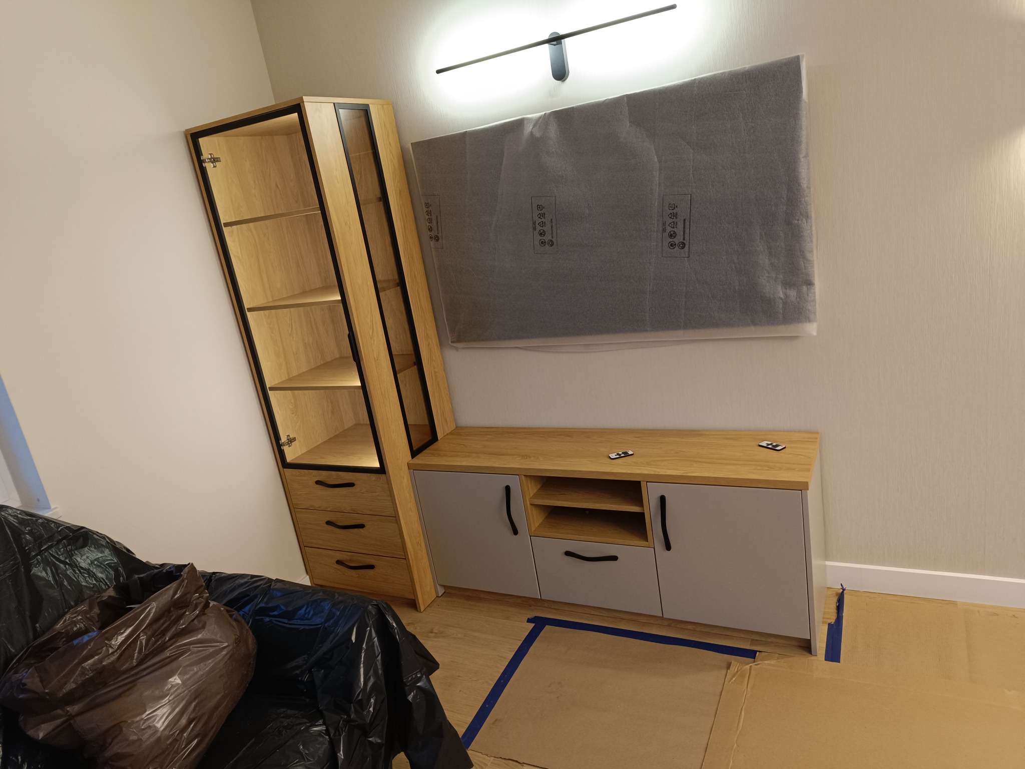 Nowoczesna meblościanka w trakcie montażu: szafka z szufladami i witryną, szara szafka RTV z drewnianym blatem, telewizor jeszcze w folii ochronnej, zabezpieczona podłoga.