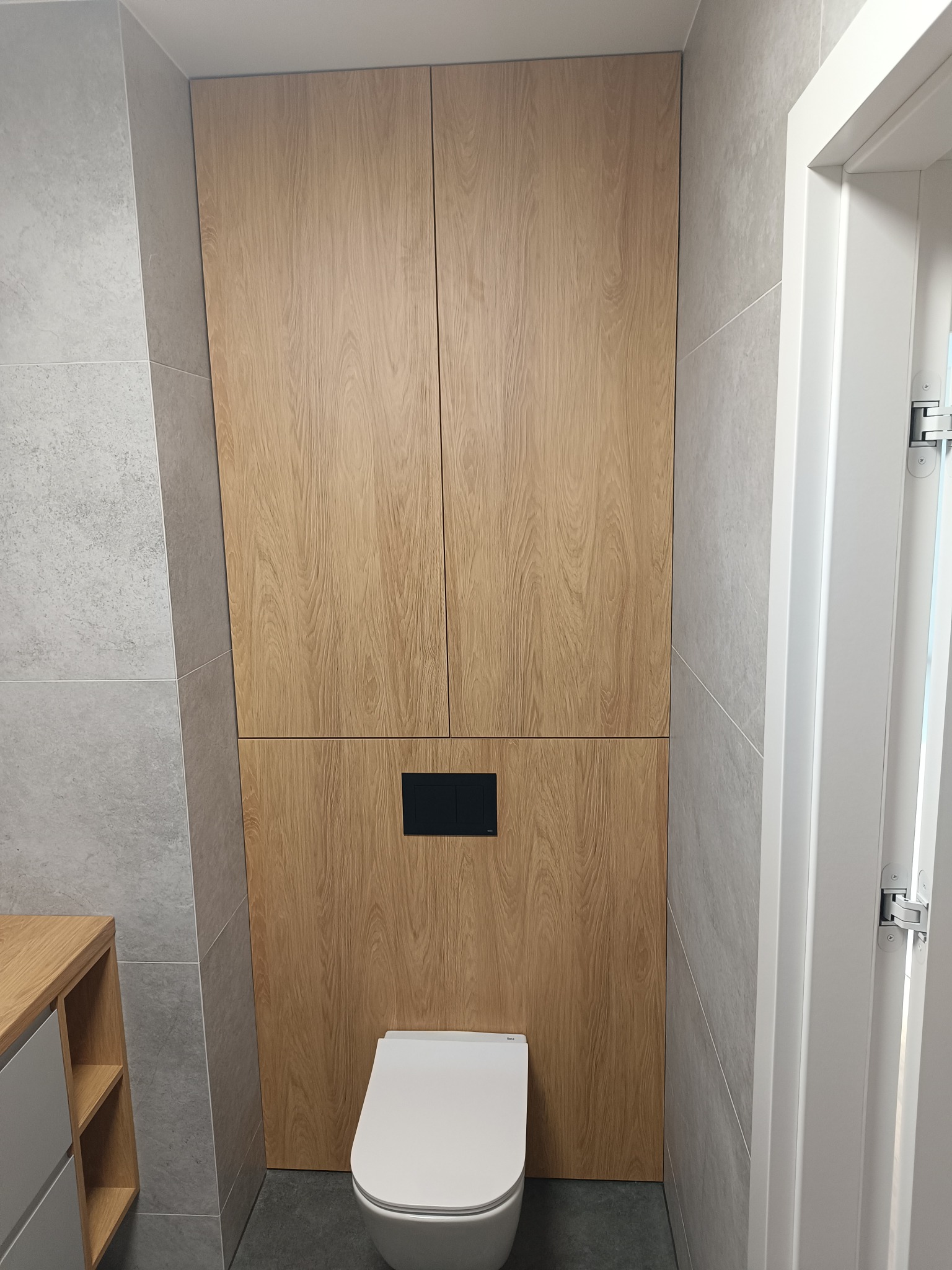 Toaleta podwieszana w zabudowie z drewnianą szafką wnękową, z czarnym panelem sterującym spłukiwaniem i szarą płytką na ścianach.