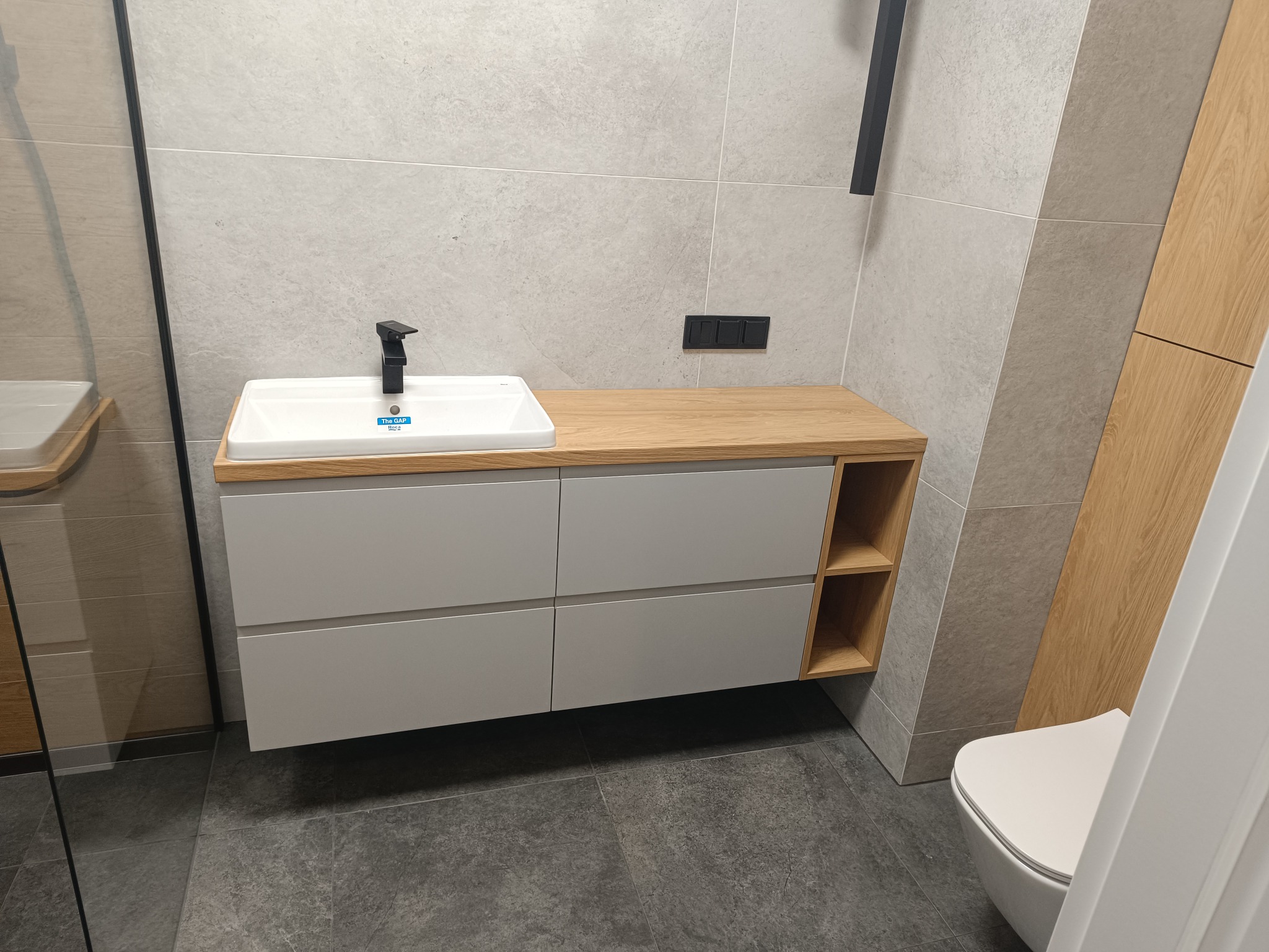 Nowoczesna łazienka z szafką pod umywalkę z szarymi frontami i blatem z jasnego drewna, czarna bateria umywalkowa, szare płytki na ścianach i podłodze.