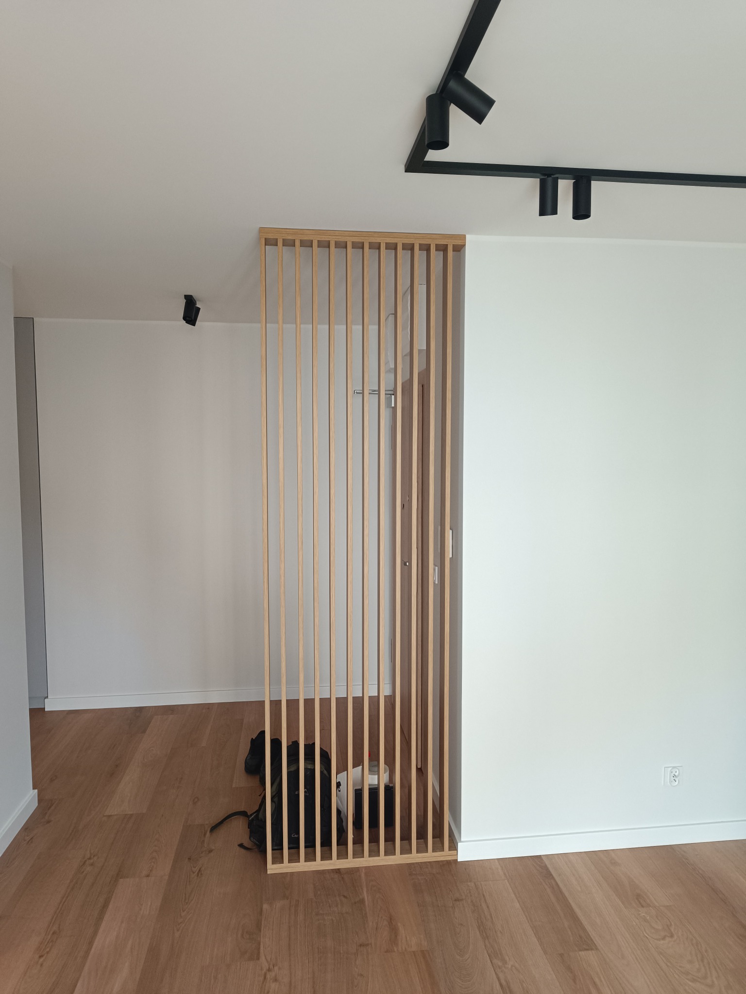 Drewniana ażurowa ścianka działowa z pionowych listew, oddzielająca przestrzeń w jasnym wnętrzu z drewnianą podłogą i nowoczesnym oświetleniem sufitowym.