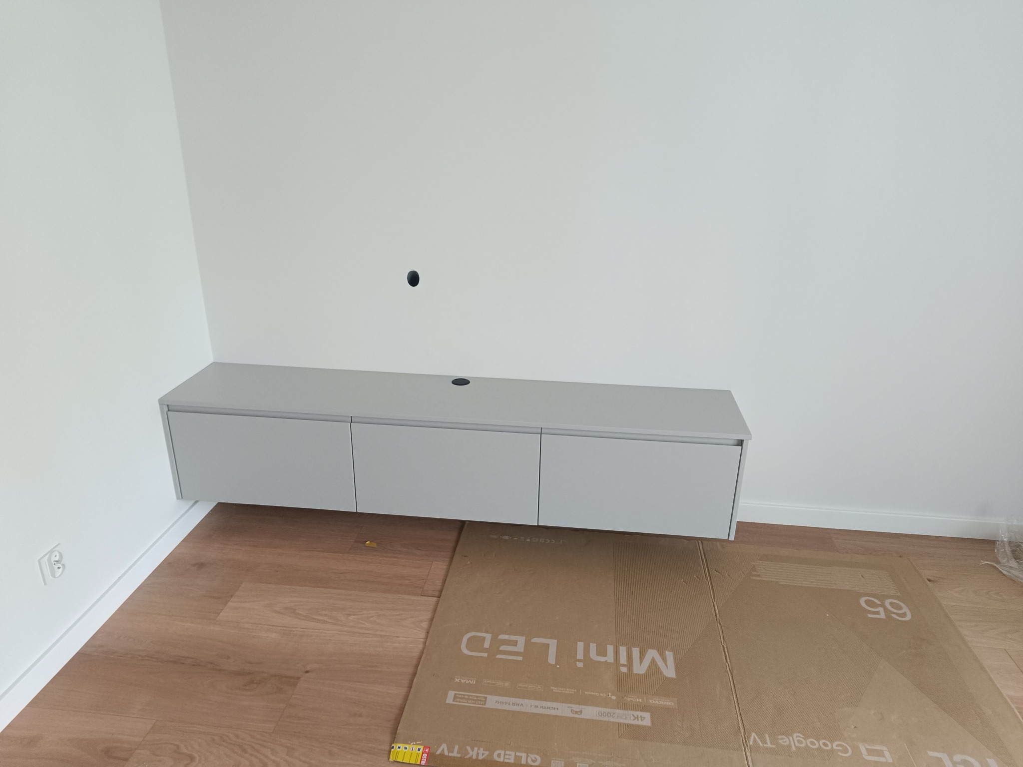 Szara szafka RTV podwieszana na ścianie, z trzema frontami, z widocznym otworem na kable, na podłodze karton po telewizorze, świeżo po montażu.