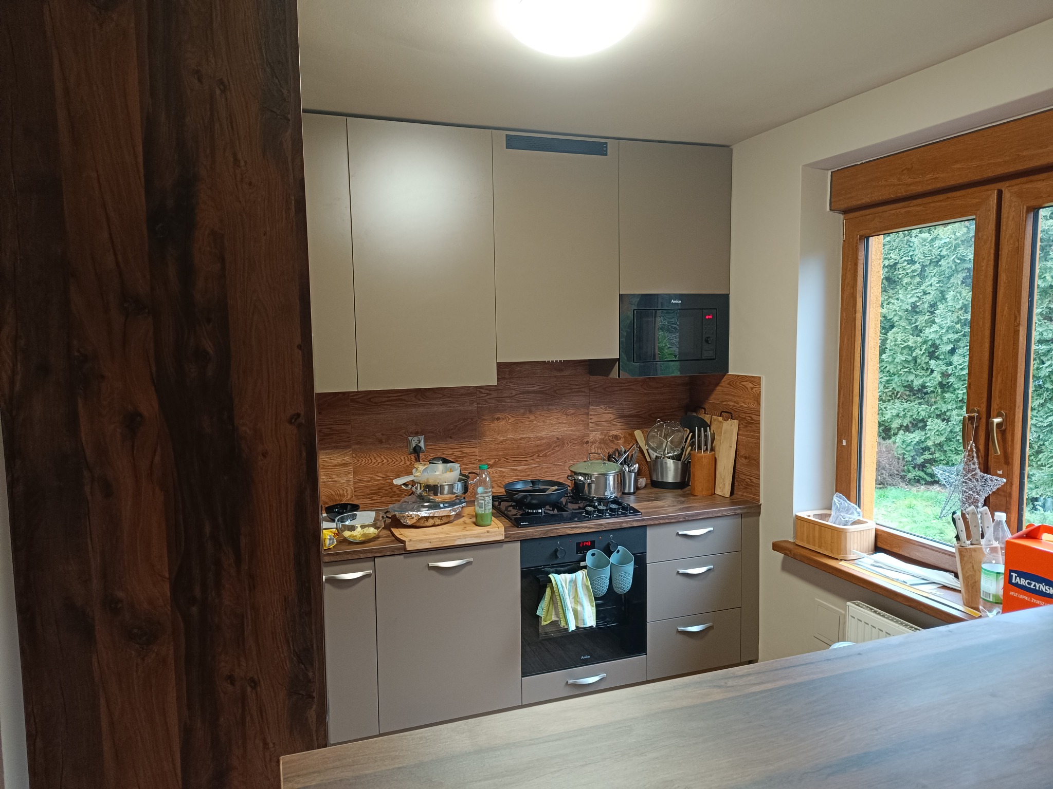 Nowoczesna kuchnia z szarymi szafkami, drewnianym blatem i panelem, czarnym piekarnikiem Amica i płytą grzewczą, z oknem z widokiem na zieleń.