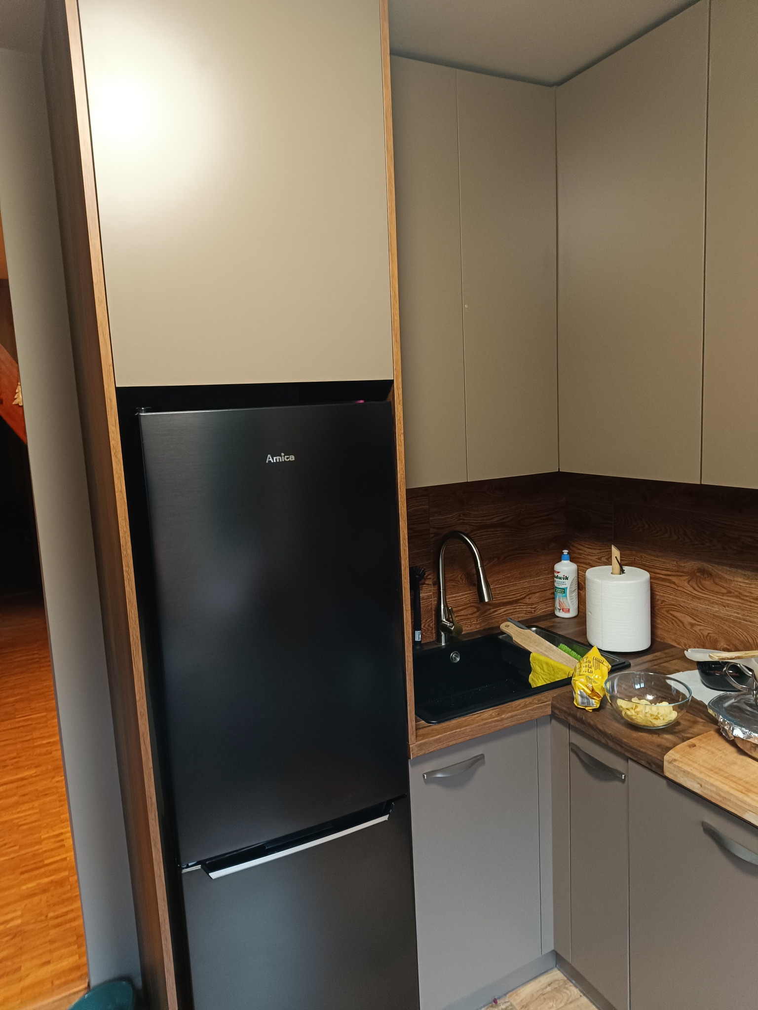 Nowoczesna kuchnia z zabudową w odcieniach beżu i szarości, z czarną lodówką Amica w zabudowie i drewnianym blatem. Na blacie widoczne akcesoria kuchenne i produkty spożywcze.