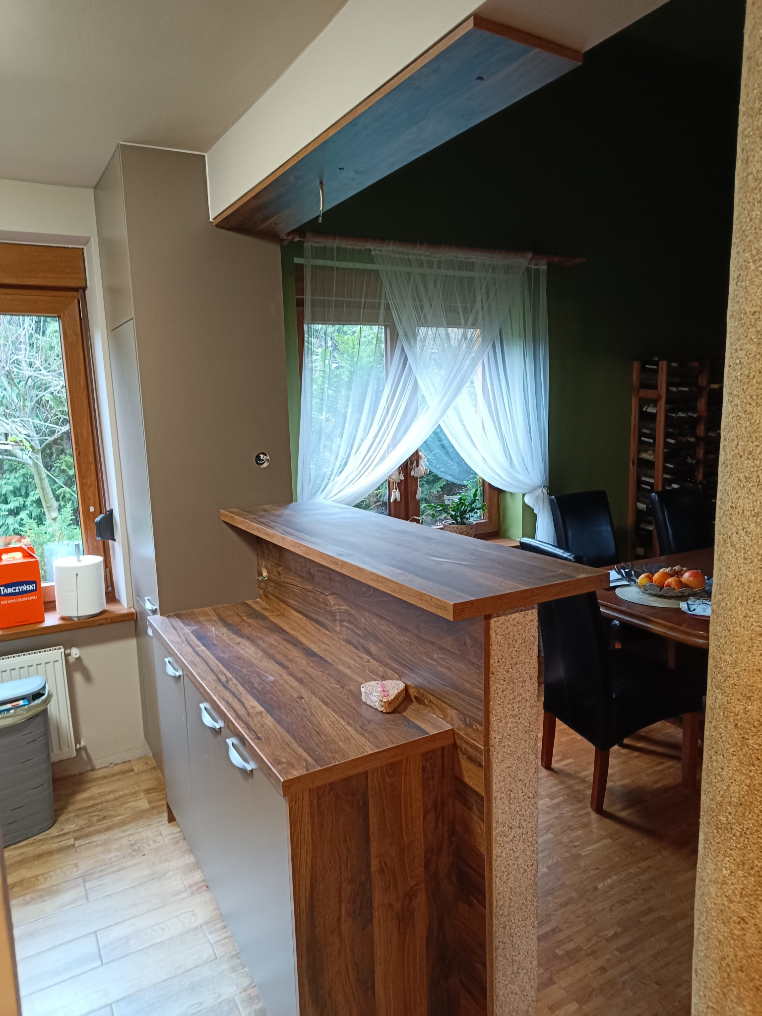 Wnętrze kuchni z półwyspem z blatem w odcieniach brązu, widok na stół z owocami i krzesła, okno z firanką w tle.