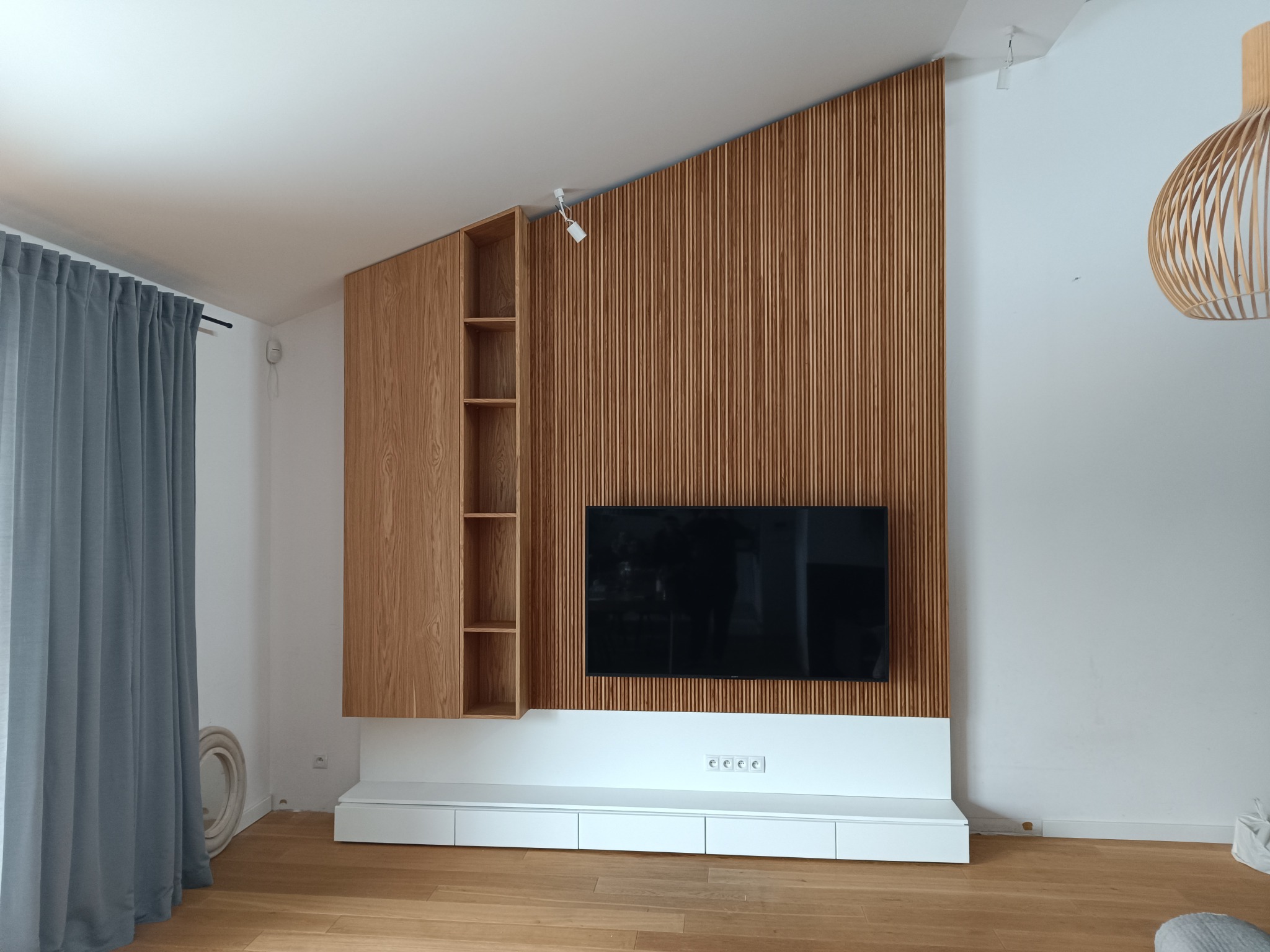 Nowoczesna ściana telewizyjna z drewnianymi lamelami, półkami i białą szafką RTV, uzupełniona wiszącą lampą z drewnianych listewek i szarą zasłoną.