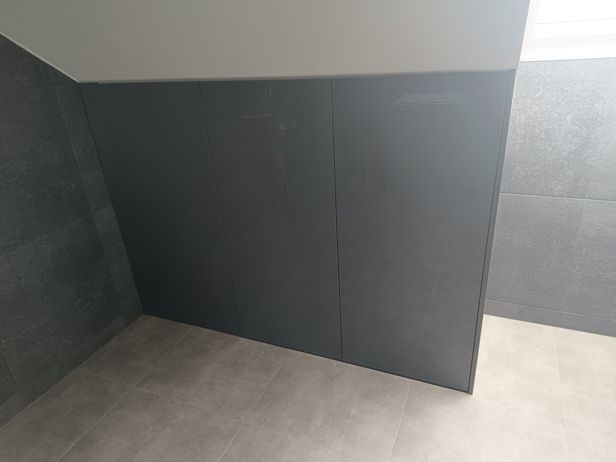Szara szafa wnękowa z połyskiem, dopasowana do skosu dachu, zintegrowana z szarą ścianą i podłogą z płytek ceramicznych.