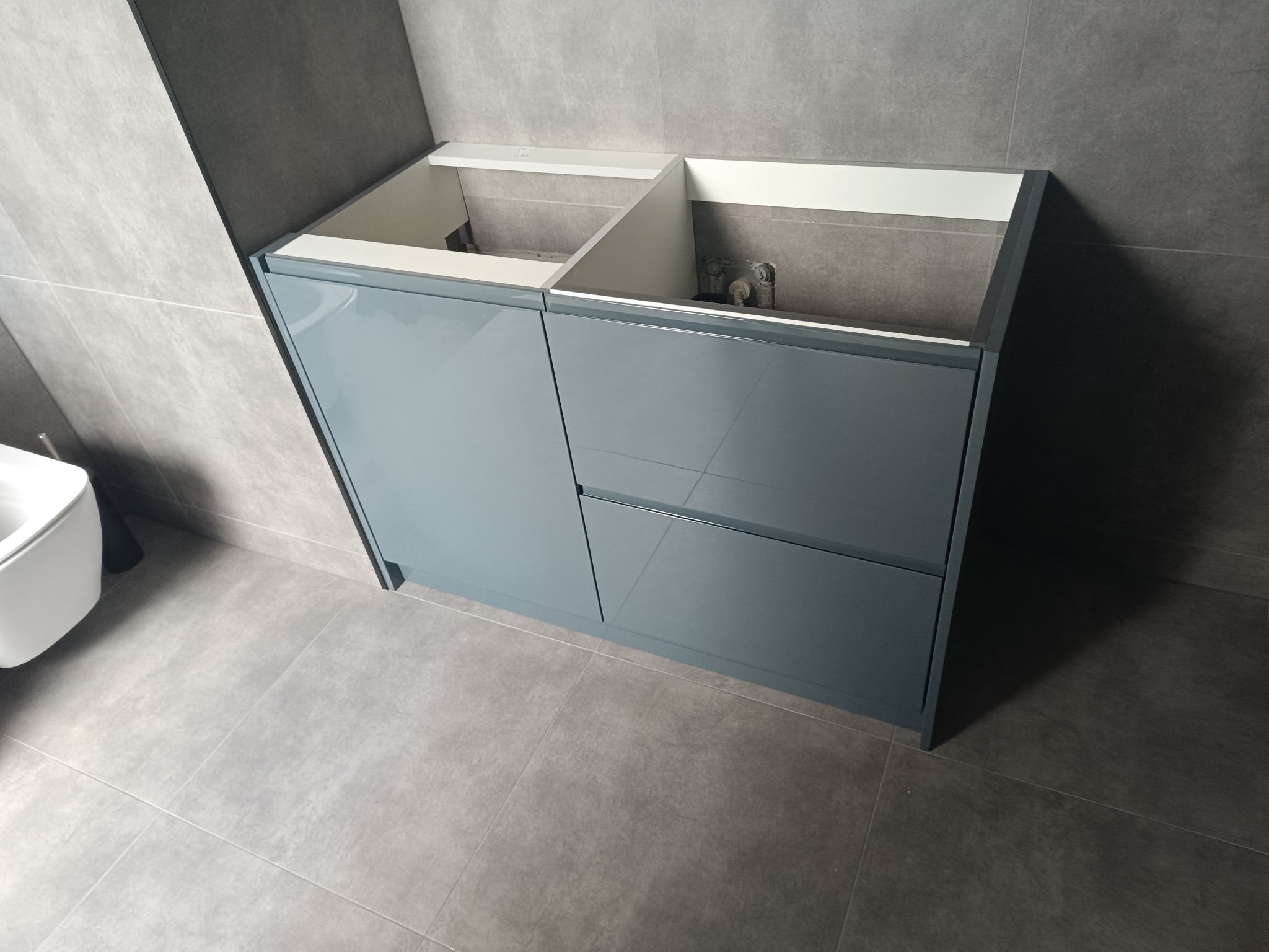 Niebieska szafka łazienkowa pod umywalkę z dwiema szufladami, bez umywalki i baterii, na tle szarych płytek ściennych i podłogowych.