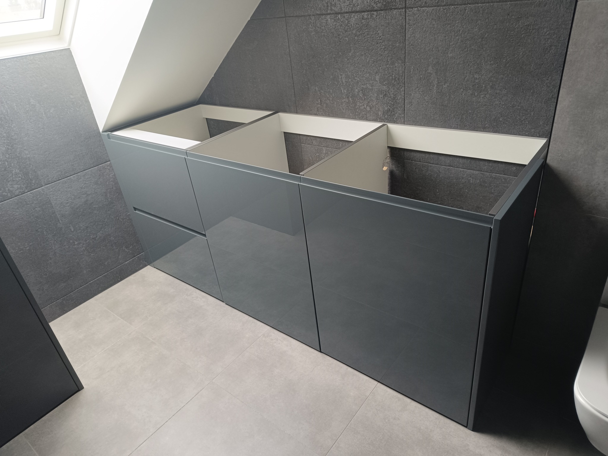 Szara szafka łazienkowa z trzema otworami na umywalki, zamontowana pod skosem dachu, z szarą płytką na ścianie i szarą podłogą.