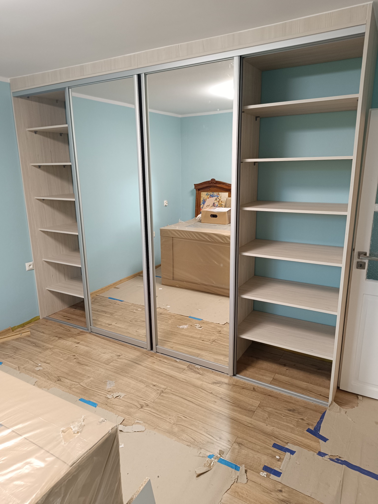 Jasna szafa wnękowa z przesuwanymi lustrzanymi drzwiami i otwartymi półkami po bokach, widoczna podczas montażu, z zabezpieczeniami na podłodze i folią ochronną na meblach w tle.