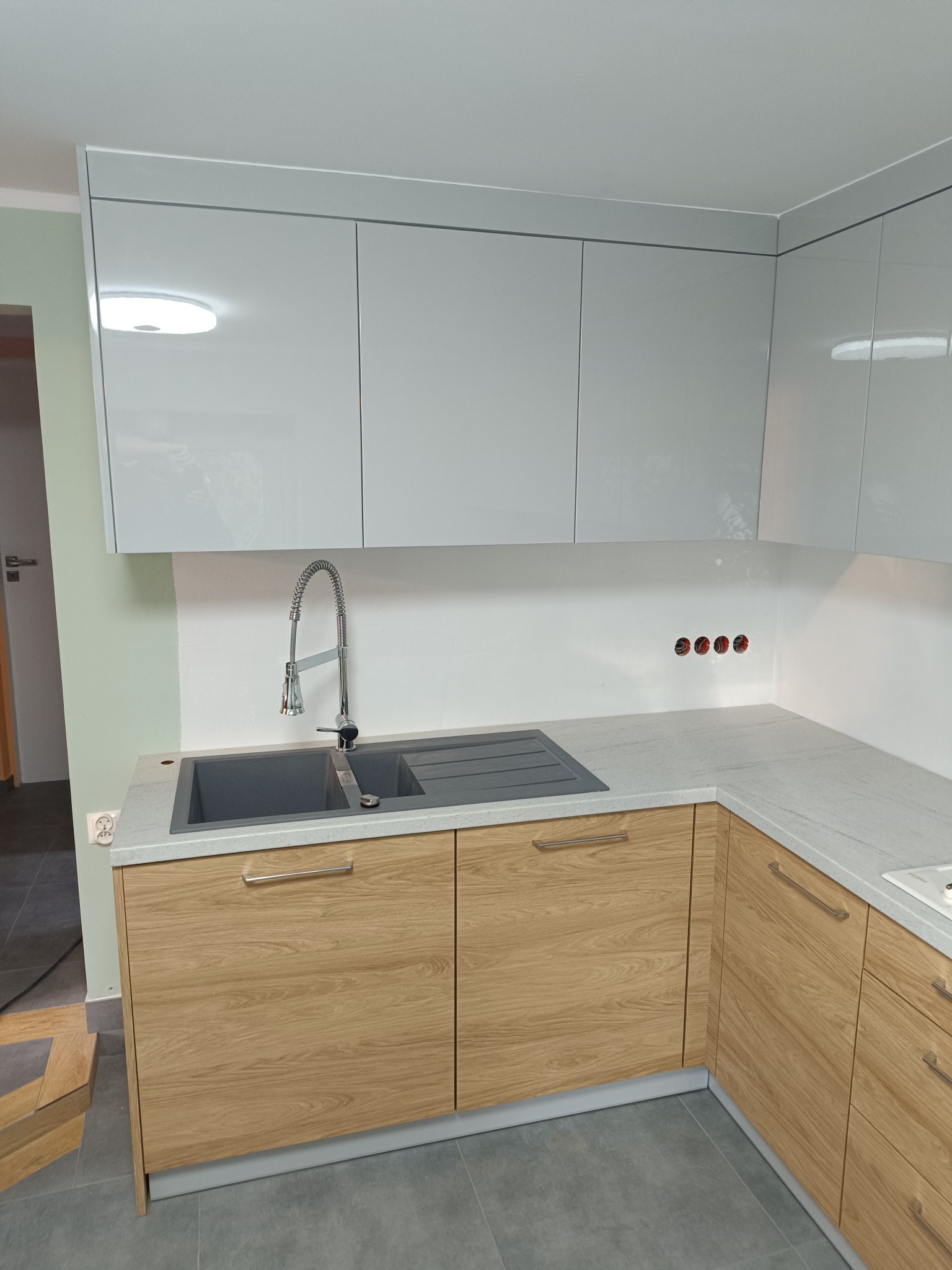 Nowoczesna kuchnia z jasnymi, lakierowanymi szafkami górnymi i drewnopodobnymi szafkami dolnymi, szarym zlewozmywakiem i baterią ze sprężyną, widoczne gniazdka elektryczne na białej ścianie.