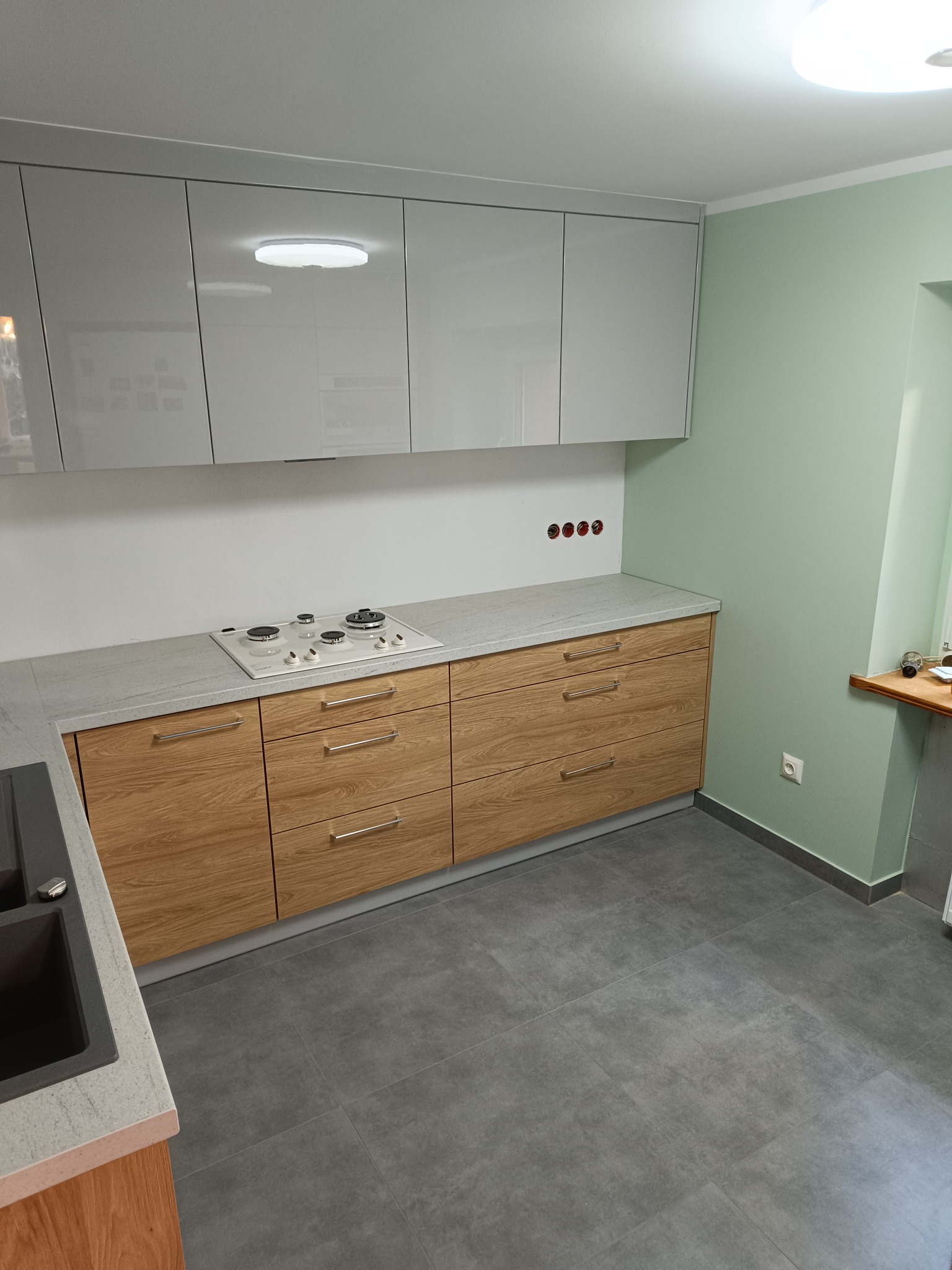 Nowoczesna kuchnia z drewnianymi frontami szafek dolnych i białymi, lakierowanymi szafkami górnymi, szary blat i płyta gazowa z białym wykończeniem.