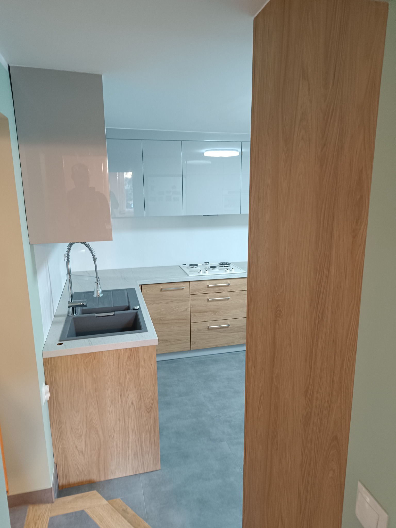 Nowoczesna kuchnia z jasnymi, lakierowanymi szafkami górnymi i drewnopodobnymi szafkami dolnymi, szarym zlewem i baterią, widoczna płyta grzewcza.