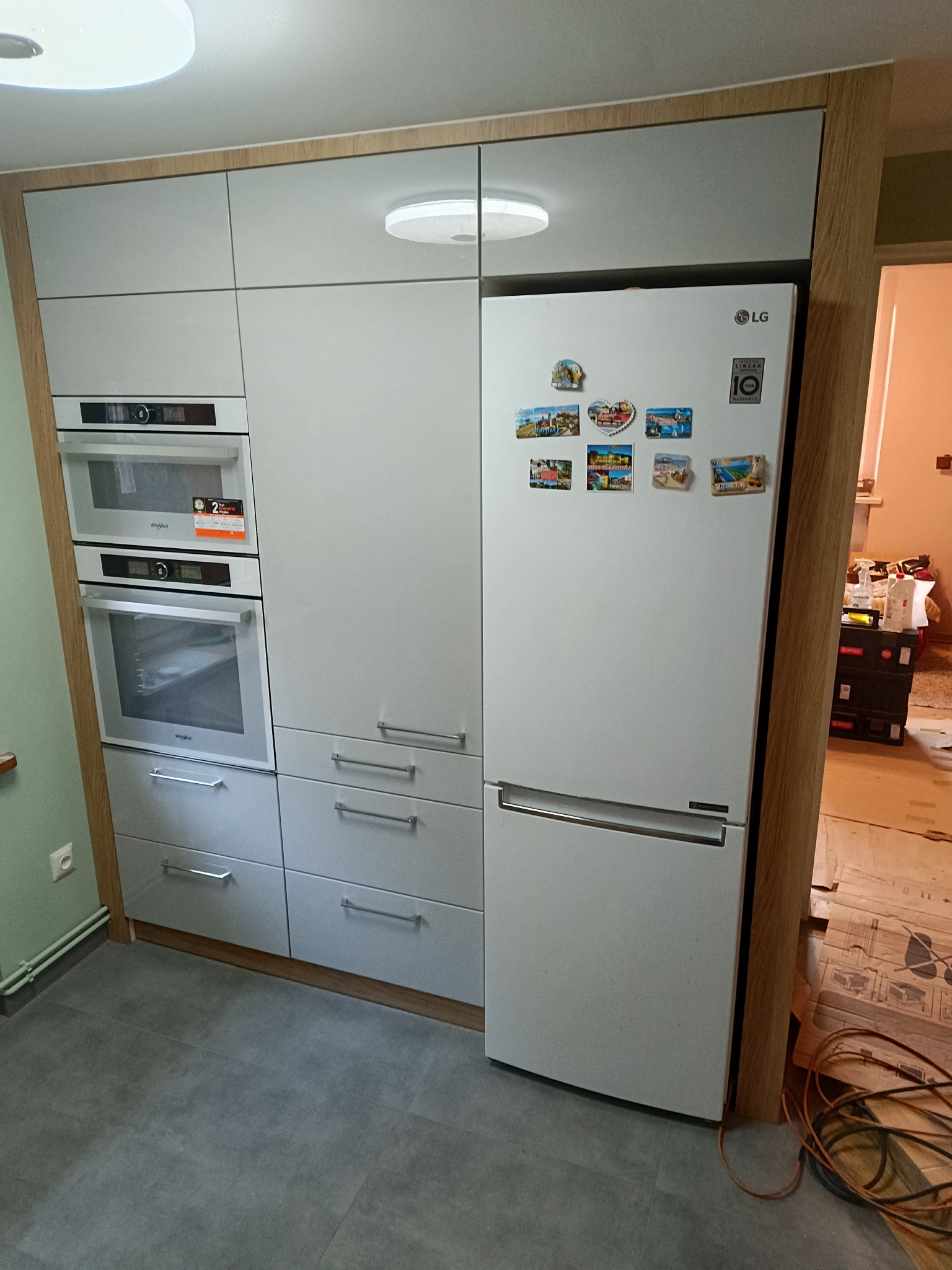 Szara zabudowa kuchenna z piekarnikami i lodówką, wykończona drewnianymi elementami, z magnesami na lodówce i widocznymi kablami na podłodze.