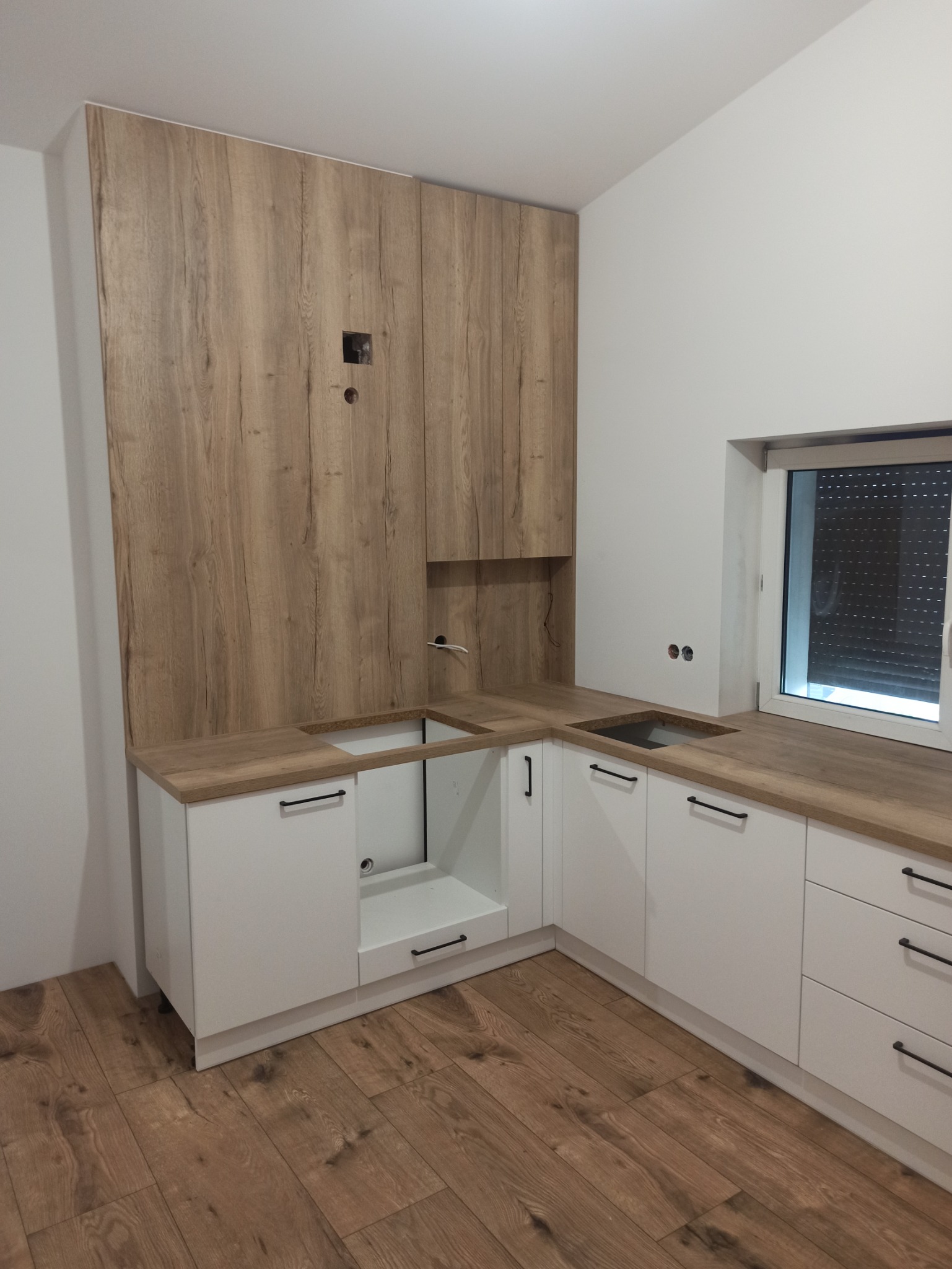 Nowoczesna, jasna kuchnia z białymi szafkami i drewnianym blatem oraz wysoką zabudową z drewnopodobną okleiną, widoczne przygotowane otwory na instalacje.