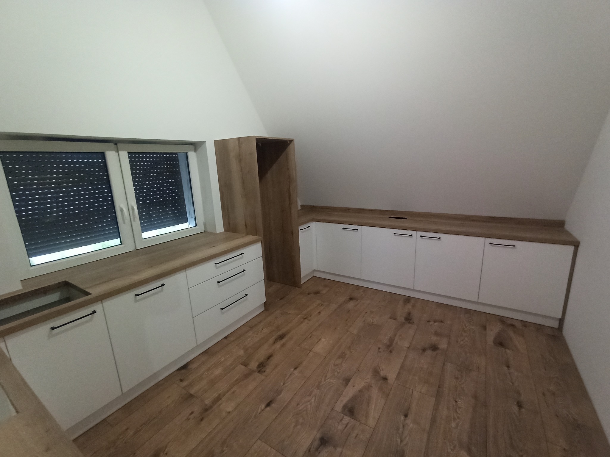 Białe szafki kuchenne z drewnianym blatem i czarnymi uchwytami w pomieszczeniu ze skośnym sufitem i drewnianą podłogą.