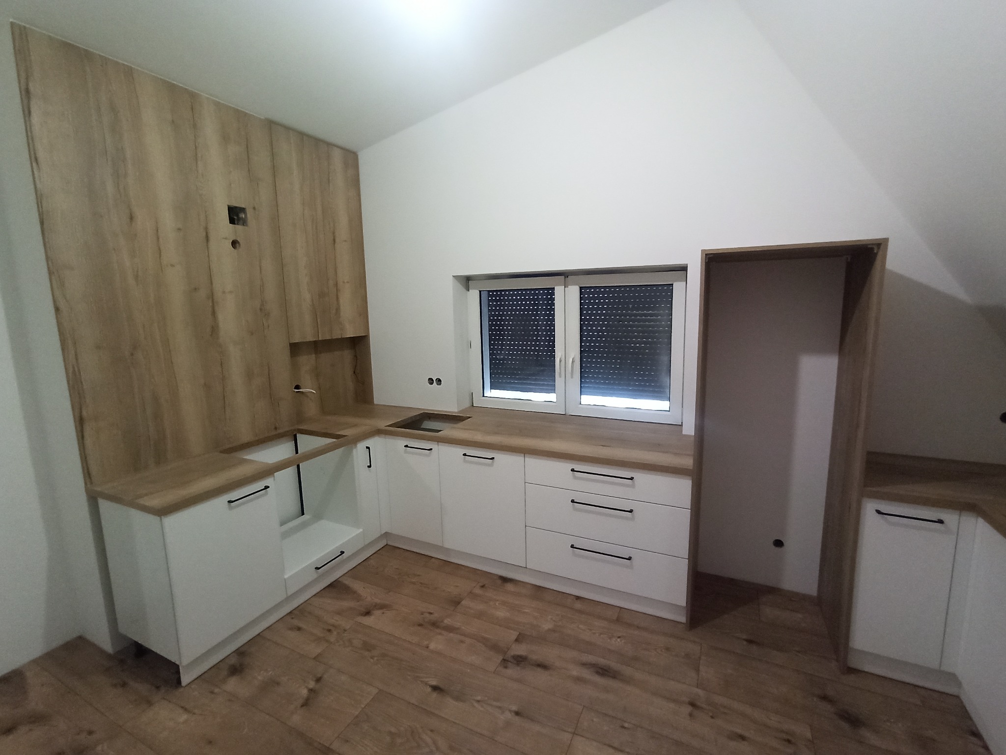 Jasna kuchnia z białymi szafkami i drewnianym blatem w pomieszczeniu ze skośnym sufitem. Drewniana okładzina ściany z widocznymi otworami na gniazdka.