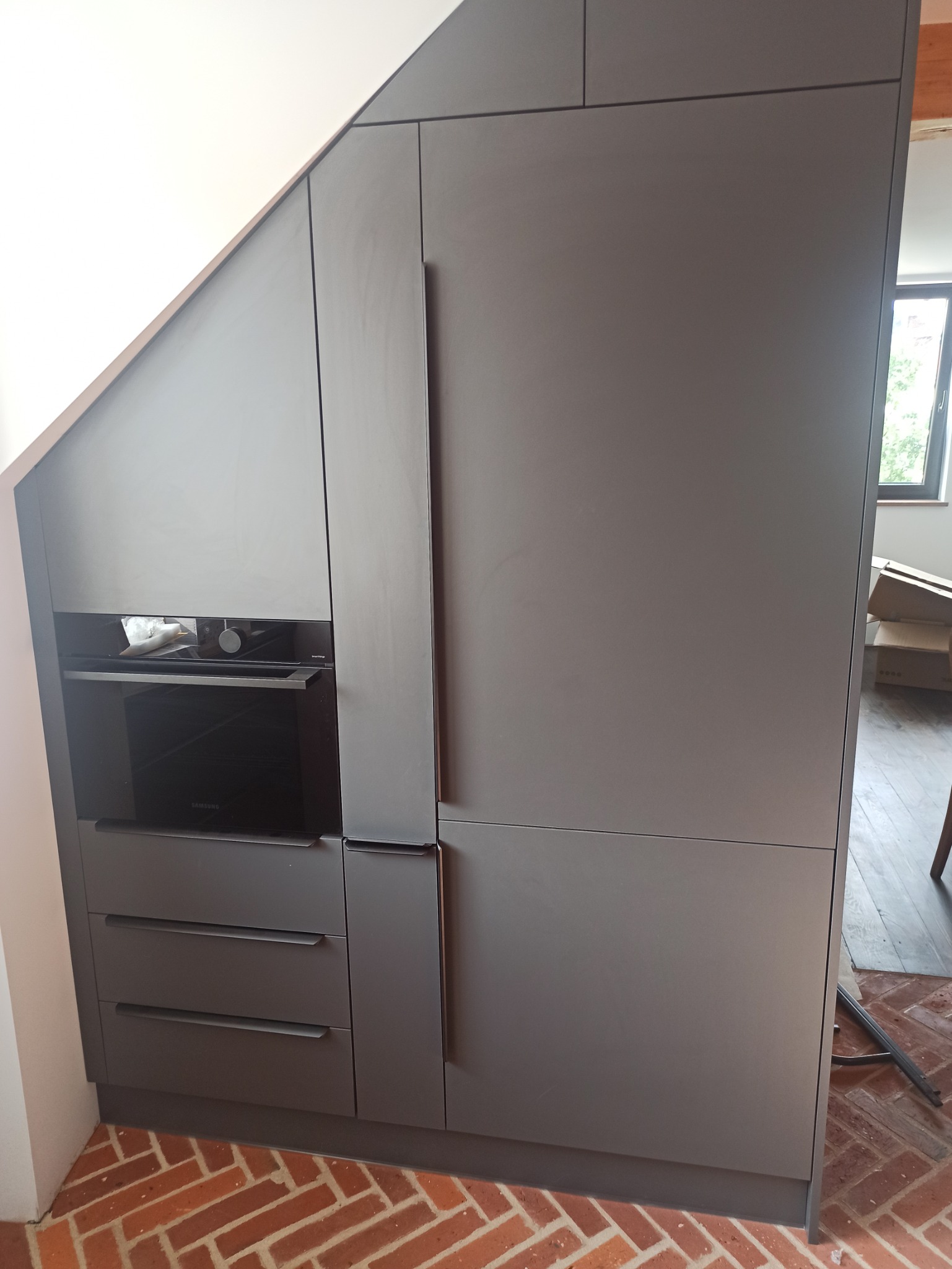 Szare szafki kuchenne na wymiar, wkomponowane w skos dachu, z piekarnikiem i szufladami, na podłodze z cegły.