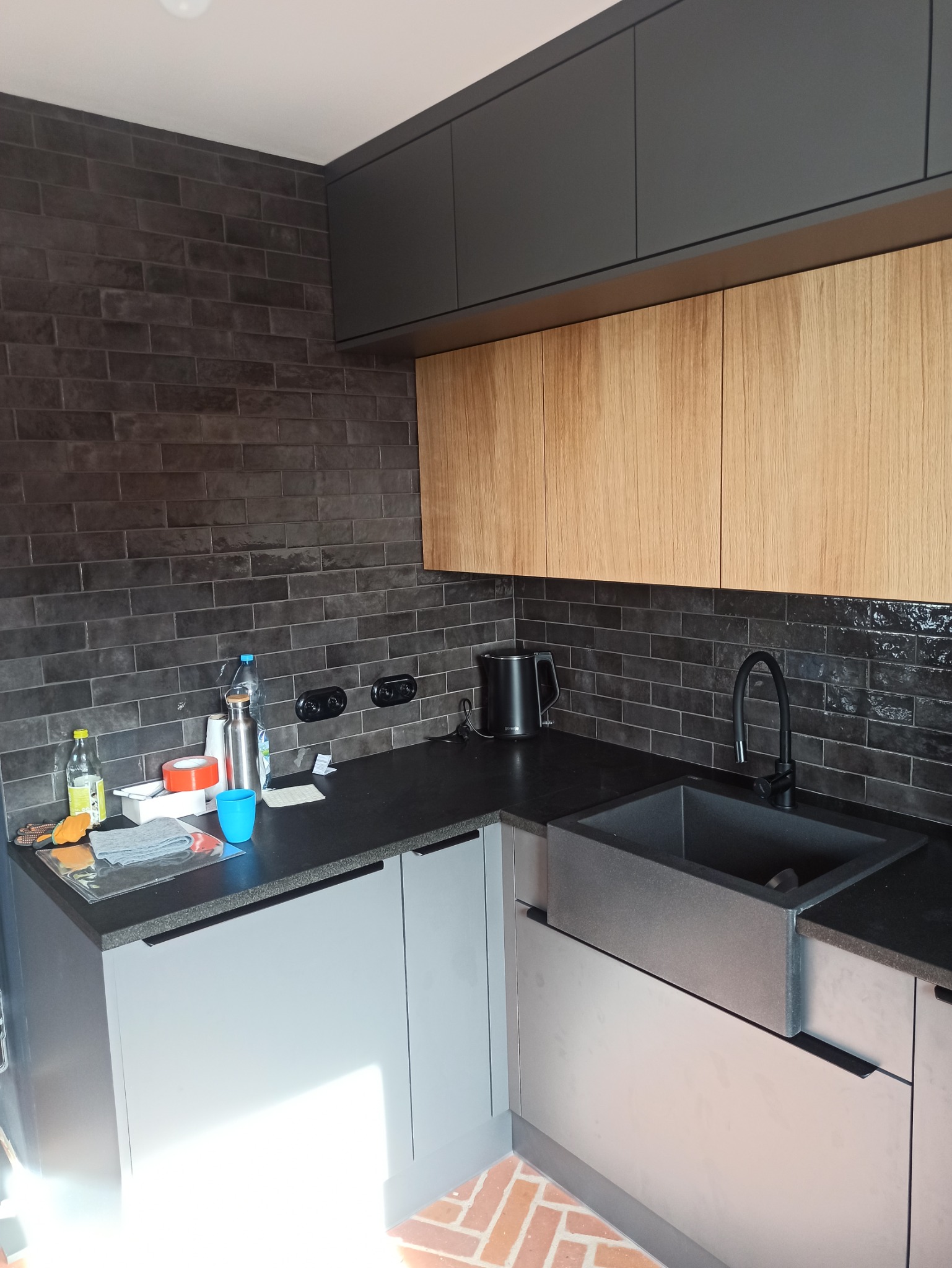 Nowoczesna kuchnia z szarymi i drewnianymi szafkami wiszącymi, czarnym blatem, ciemną ceglaną ścianą i kamiennym zlewozmywakiem.