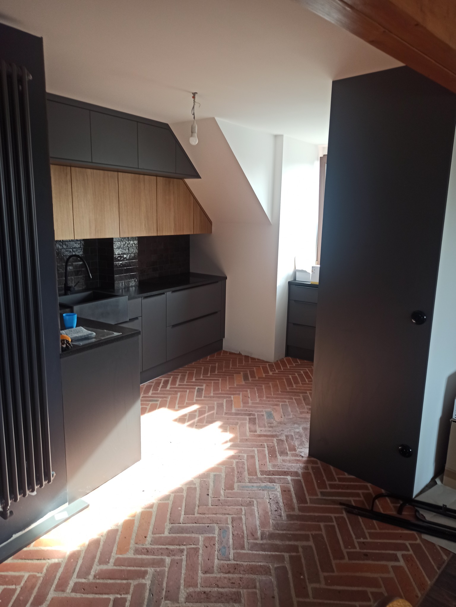 Nowoczesna kuchnia z ciemnymi szafkami, drewnianymi frontami górnych szafek i ceglaną podłogą w jodełkę, oświetlona naturalnym światłem wpadającym przez okno.