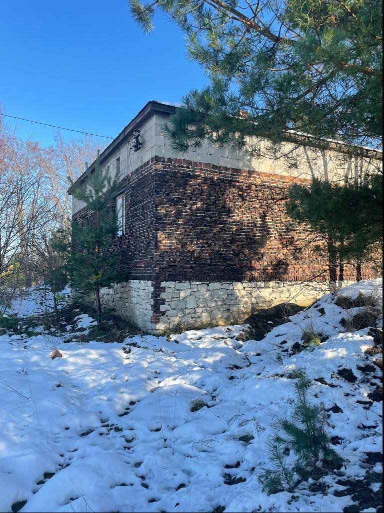 Dwukondygnacyjny budynek z cegły i kamienia w zimowej scenerii, częściowo zasłonięty przez drzewa iglaste.