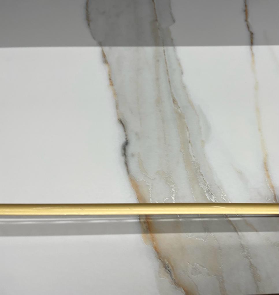 Biała płytka ceramiczna imitująca marmur z wyraźnym użyleniem w odcieniach szarości i złota, uzupełniona o poziomy, złoty reling łazienkowy.