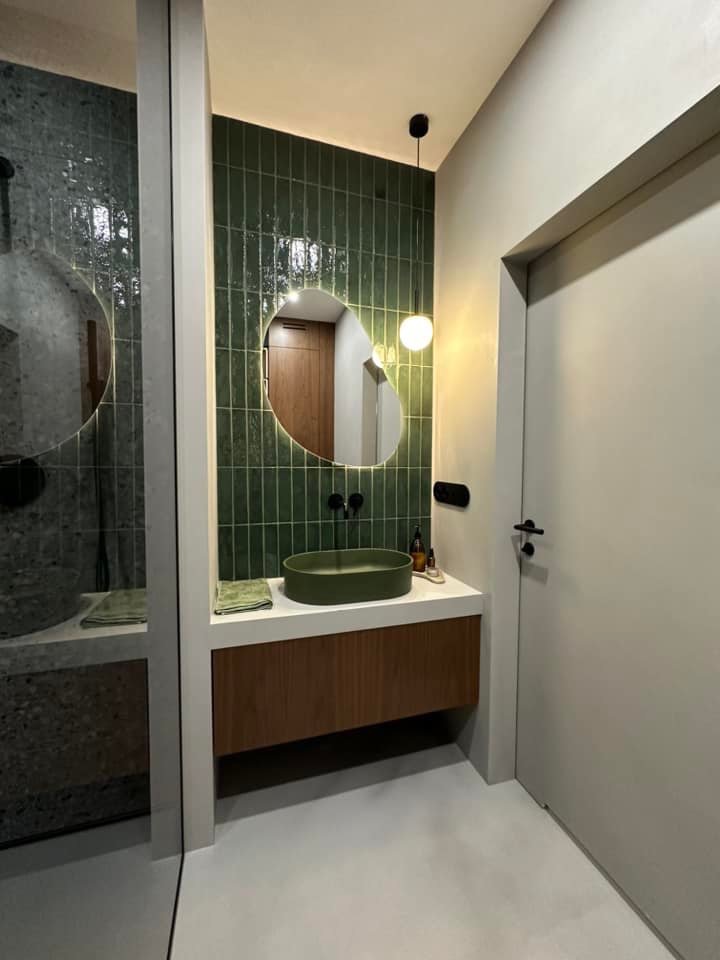 Nowoczesna łazienka z zielonymi płytkami ułożonymi pionowo, owalną umywalką w tym samym odcieniu, podświetlanym lustrem o nieregularnym kształcie i drewnianą szafką pod umywalką.