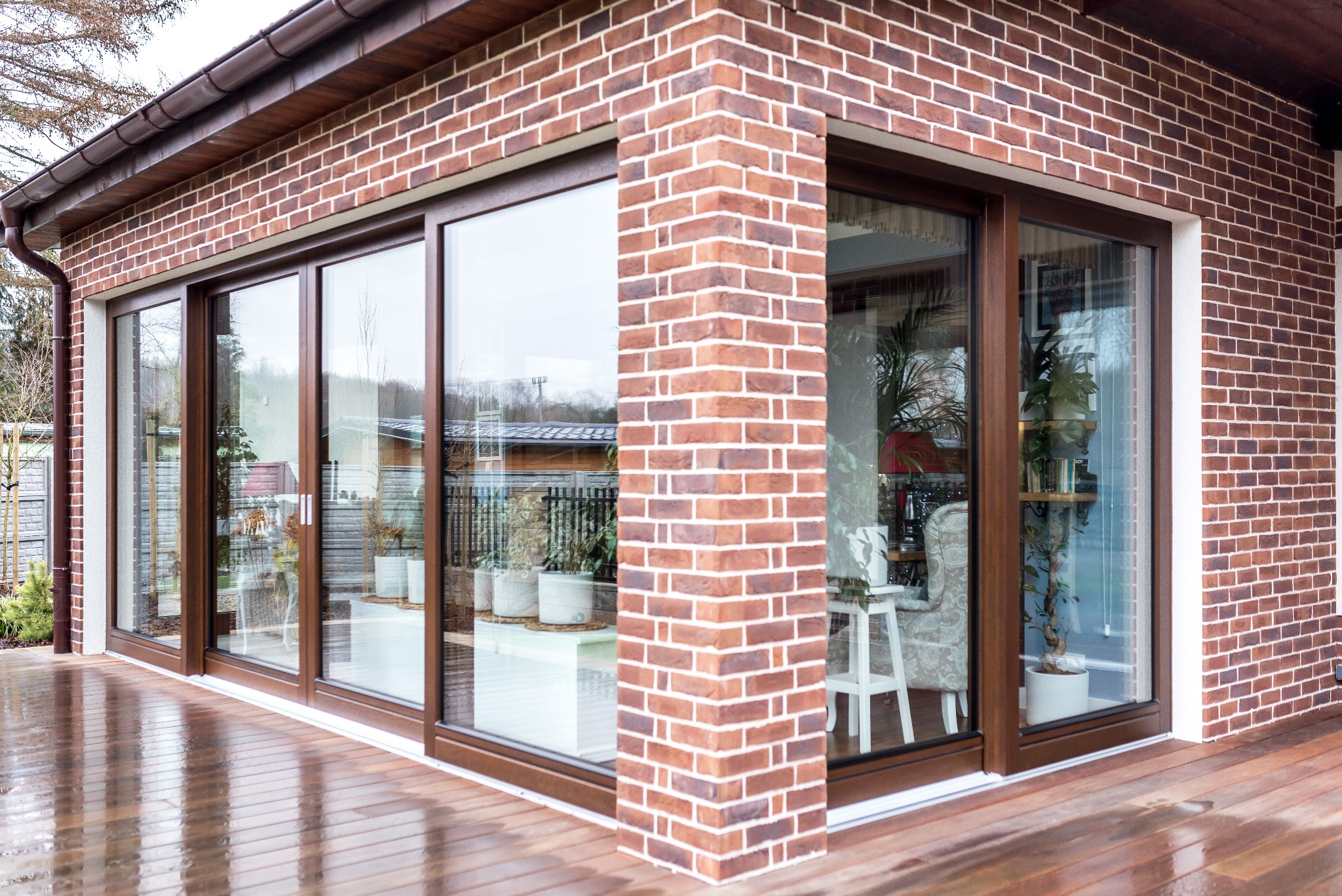 Nowoczesny dom z czerwonej cegły z dużymi przesuwnymi oknami z brązowymi ramami, widoczne odbicia na drewnianym tarasie po deszczu.