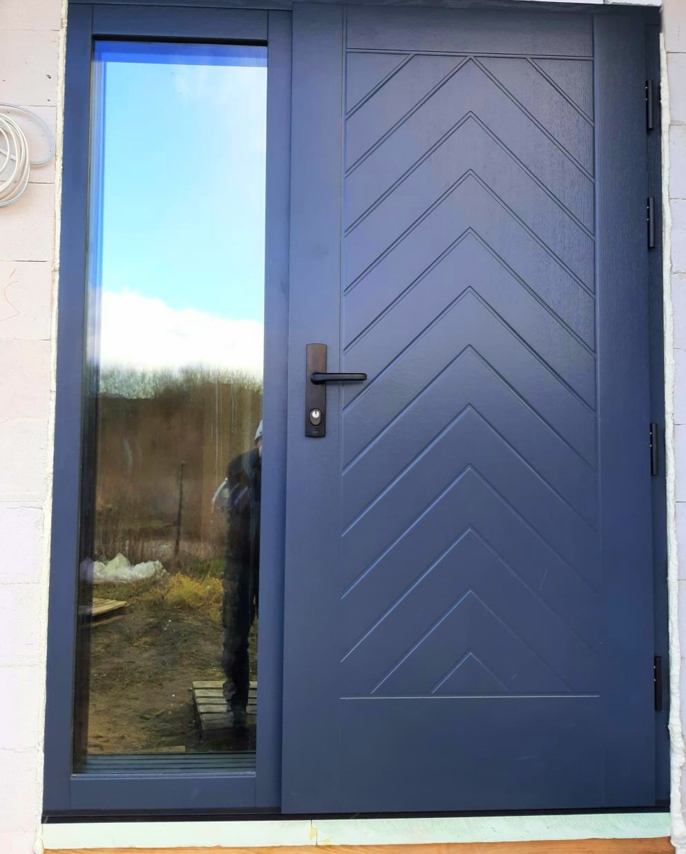 Nowoczesne, grafitowe drzwi zewnętrzne z bocznym przeszkleniem, z wzorem jodełki na skrzydle, zamontowane w surowym murze.