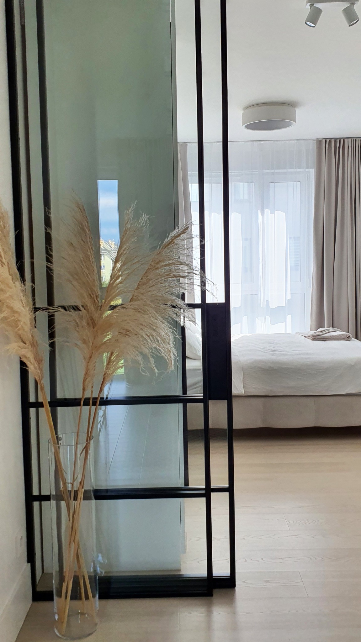 Szklane drzwi przesuwne z czarną ramą w sypialni, widok na łóżko i okno z firankami, dekoracja z trawy pampasowej w wazonie.