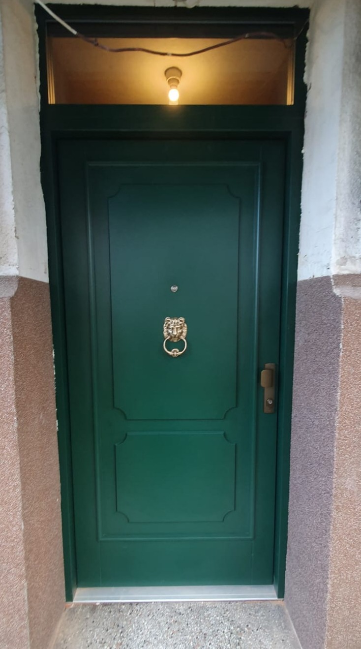 Zielone drzwi wejściowe z ozdobną kołatką w kształcie lwa, zamontowane w starej kamienicy, oświetlone od góry żarówką.