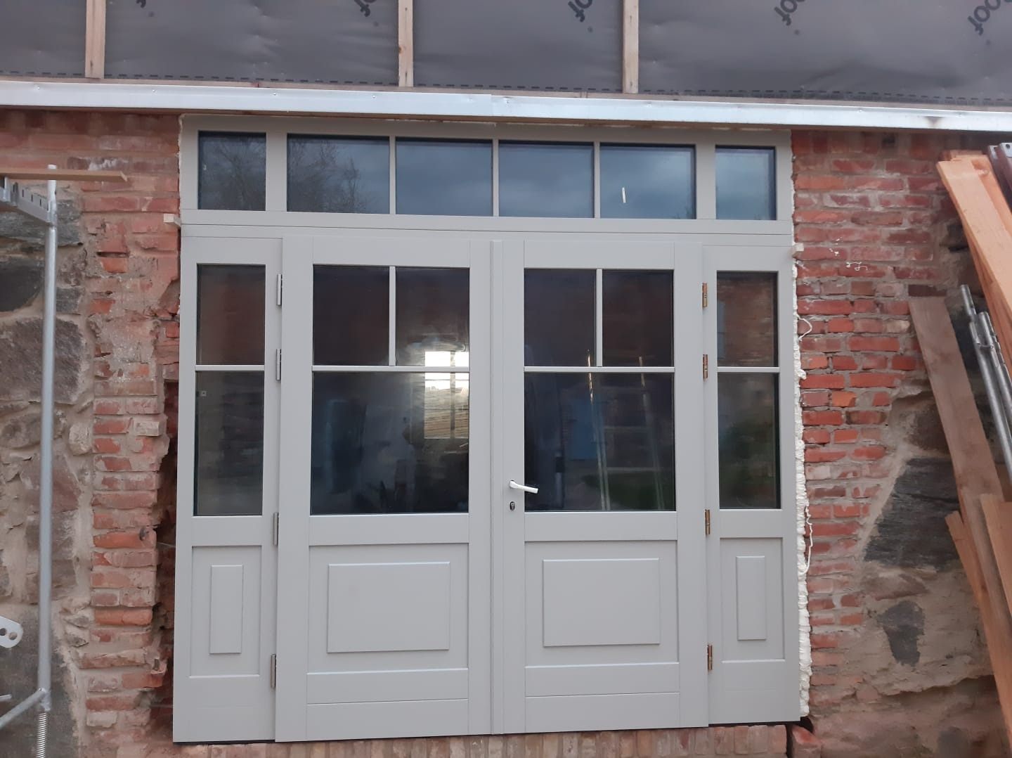 Szare drzwi dwuskrzydłowe z nadświetlem zamontowane w surowej, ceglanej ścianie podczas remontu, widoczne pianka montażowa i rusztowanie.