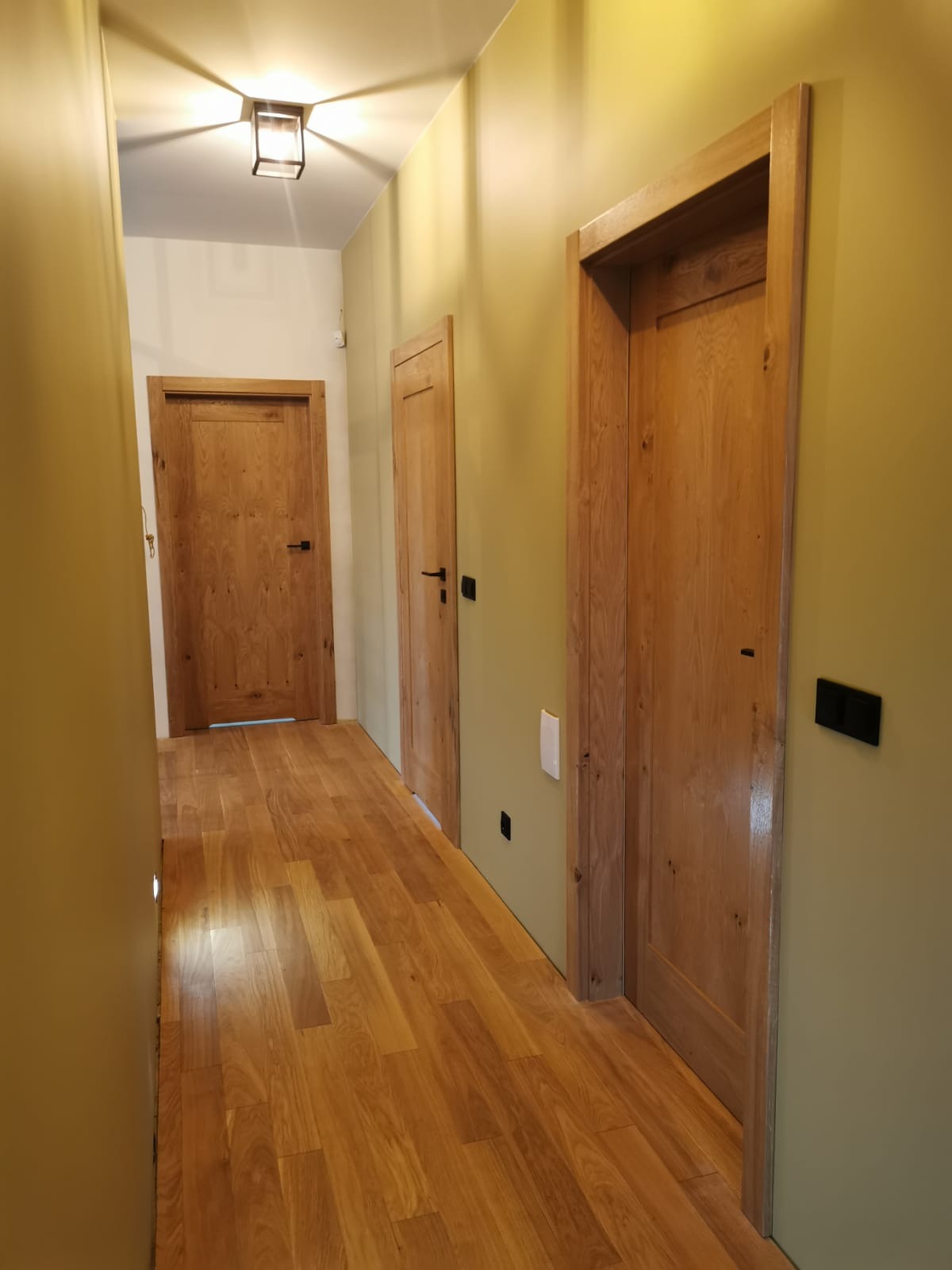 Korytarz z trzema drewnianymi drzwiami wewnętrznymi o naturalnym odcieniu drewna, widok z perspektywy podłogi z parkietu w kierunku oświetlonego sufitu z lampą.