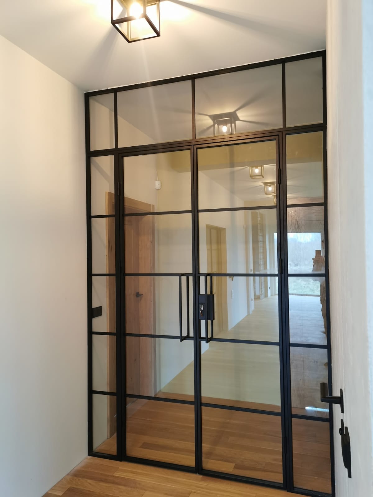 Szklane drzwi dwuskrzydłowe w czarnej, metalowej ramie z widocznymi klamkami i zamkiem, oddzielające korytarz z drewnianą podłogą i oświetleniem sufitowym.