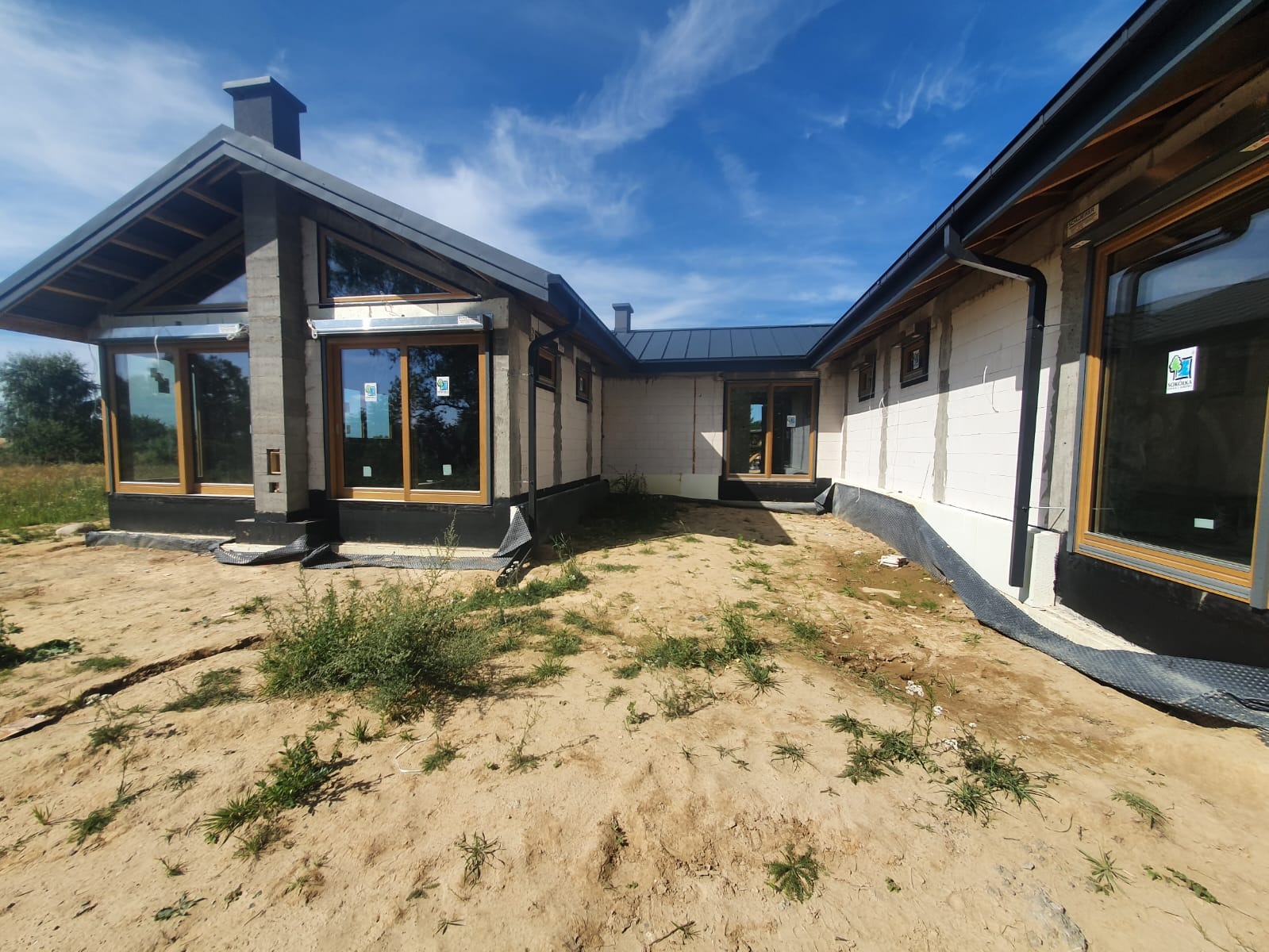 Surowy stan domu jednorodzinnego z zamontowanymi oknami z drewnianą ramą i czarną membraną przy fundamentach, widoczne bloczki betonowe ścian i niezagospodarowany teren wokół.