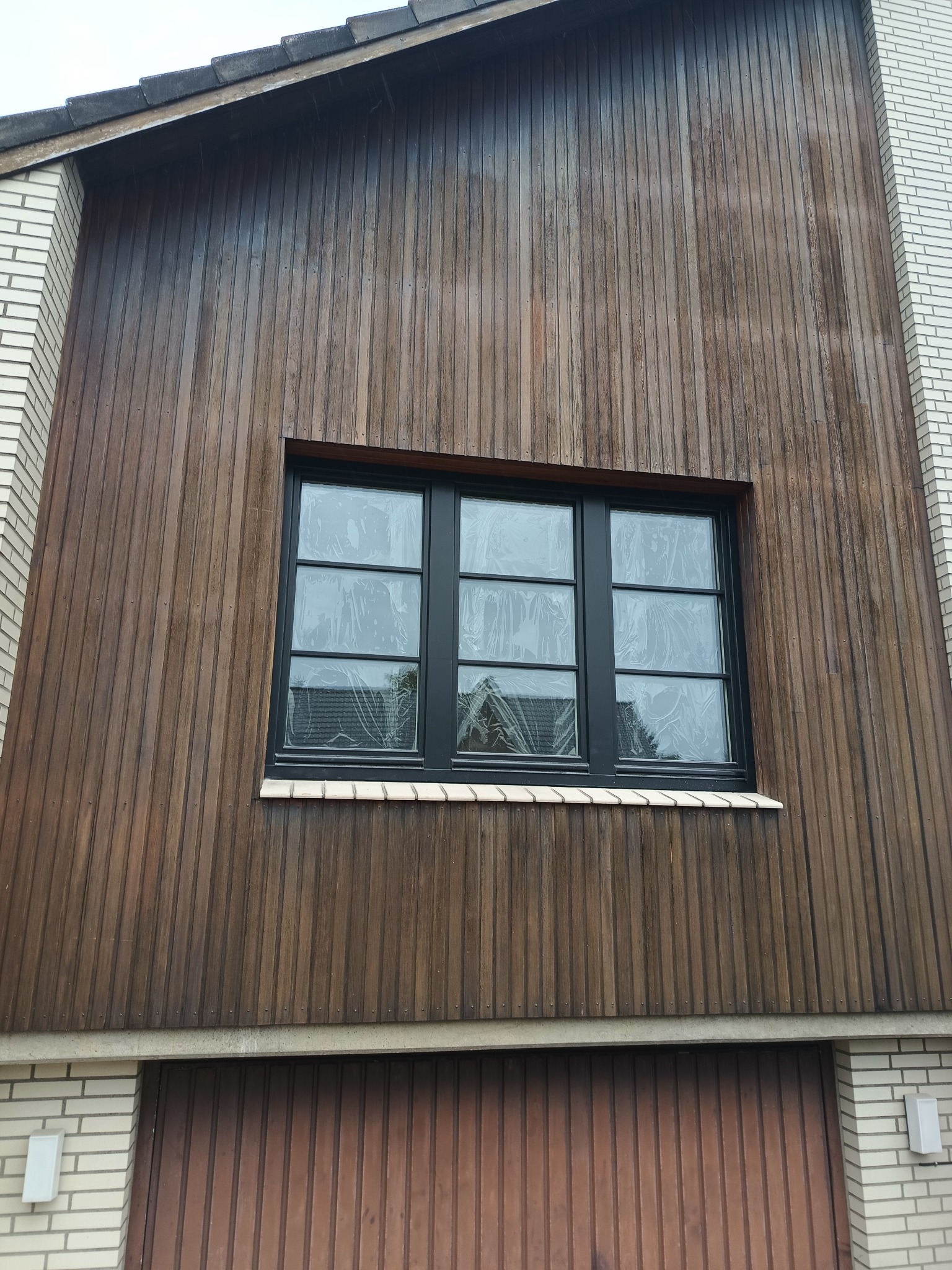 Nowe, czarne okno z podziałami szprosowymi, zabezpieczone folią ochronną, zamontowane w elewacji z drewnianych paneli i cegły klinkierowej.