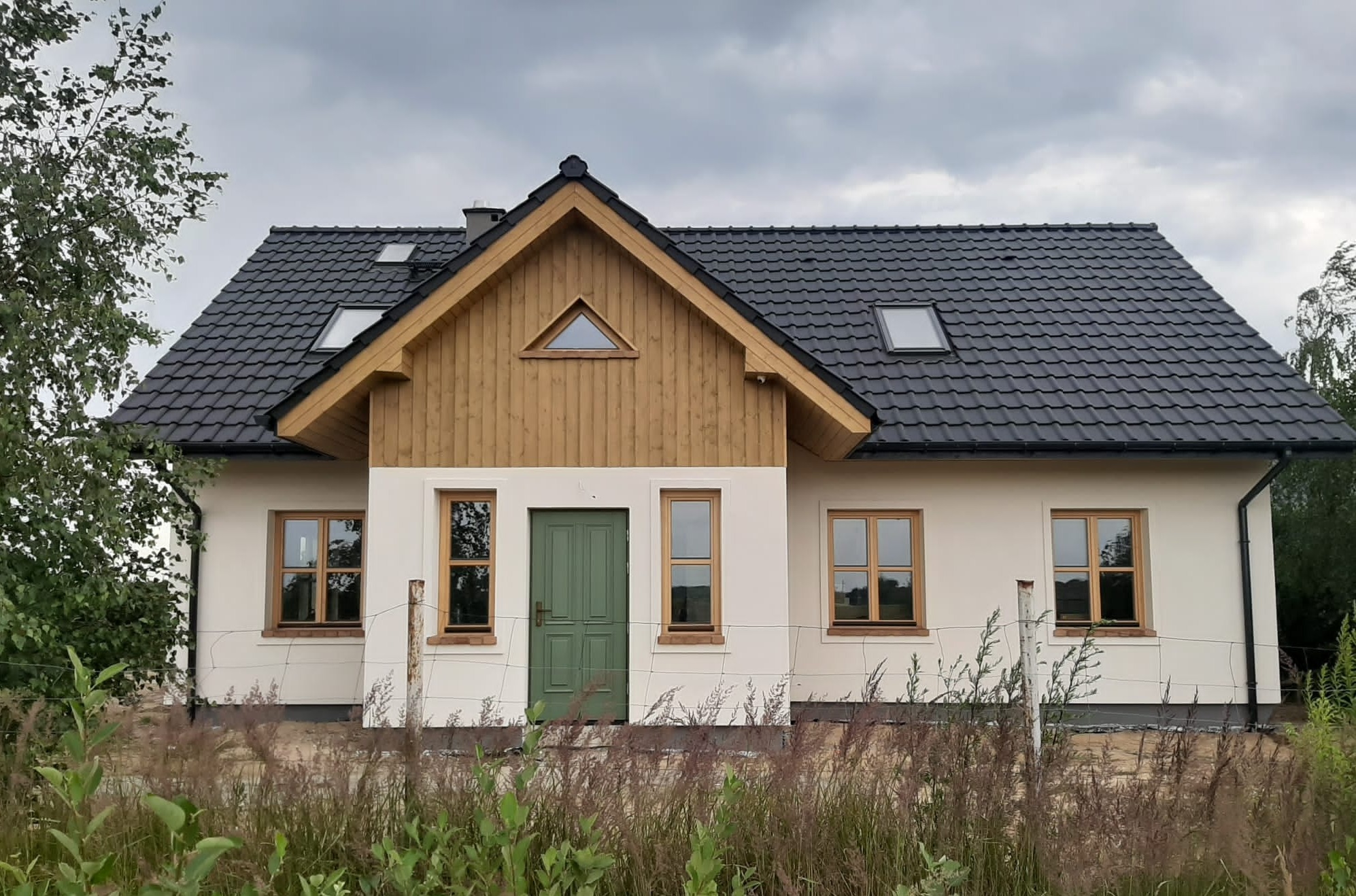 Nowo wybudowany dom jednorodzinny z ciemną dachówką, drewnianą fasadą szczytową i zielonymi drzwiami wejściowymi, widoczne drewniane ramy okienne i niezagospodarowany teren przed budynkiem.