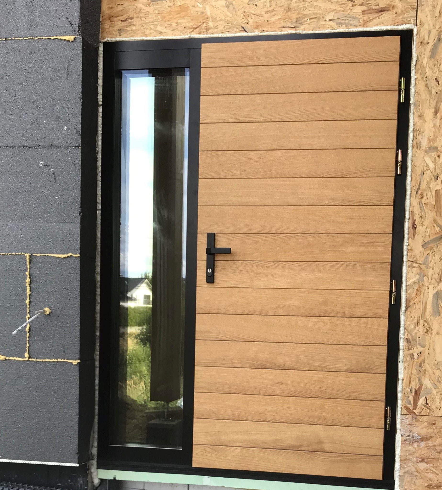 Nowoczesne drzwi zewnętrzne z panelem drewnopodobnym i szybą, czarna rama, widok z zewnątrz na surową ścianę i budynek w tle.