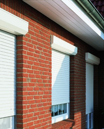 Białe rolety zewnętrzne zamontowane na ceglanej ścianie budynku, częściowo opuszczone, widoczne okna.
