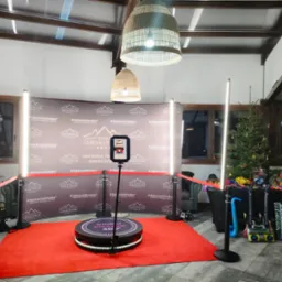 Oświetlona platforma obrotowa Videofrajda 360 na czerwonym dywanie, otoczona dekoracjami świątecznymi i rekwizytami do zdjęć, ustawiona na tle ściany z logo Osada Górski Potok.