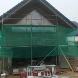 Dom w trakcie ocieplania, widoczne rusztowanie z zieloną siatką, materiały budowlane i narzędzia na placu budowy.