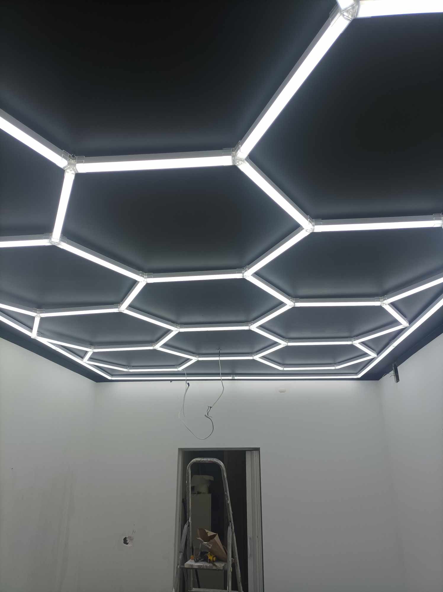 Sufit podwieszany z czarną powierzchnią i sześciokątnym oświetleniem LED, widoczna drabina i kable zwisające w surowym wnętrzu.