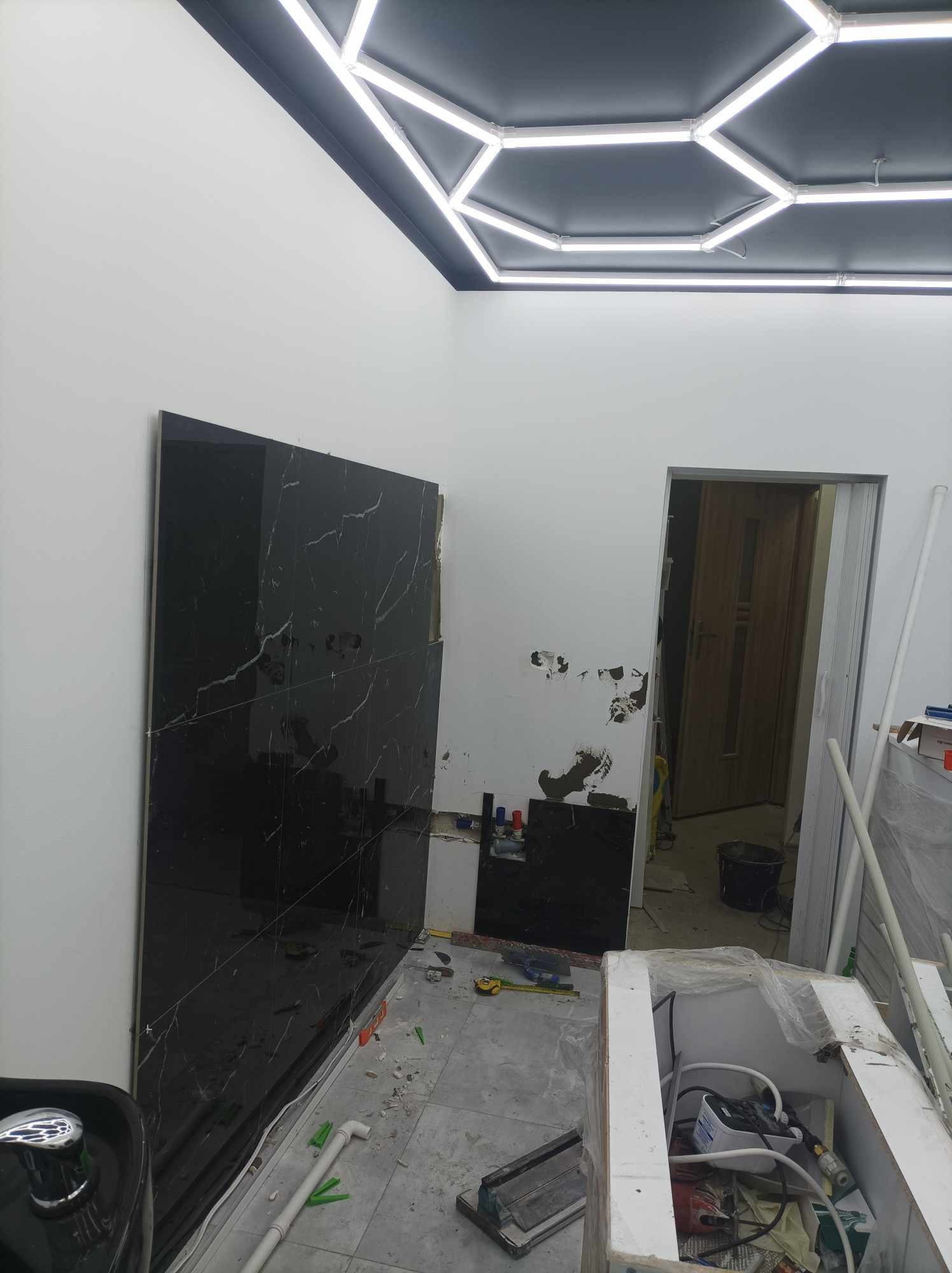 Montaż czarnych płytek imitujących marmur na ścianie w łazience w trakcie prac, widoczne narzędzia, poziomica, klej i fragment sufitu z ledowym oświetleniem w kształcie heksagonów.