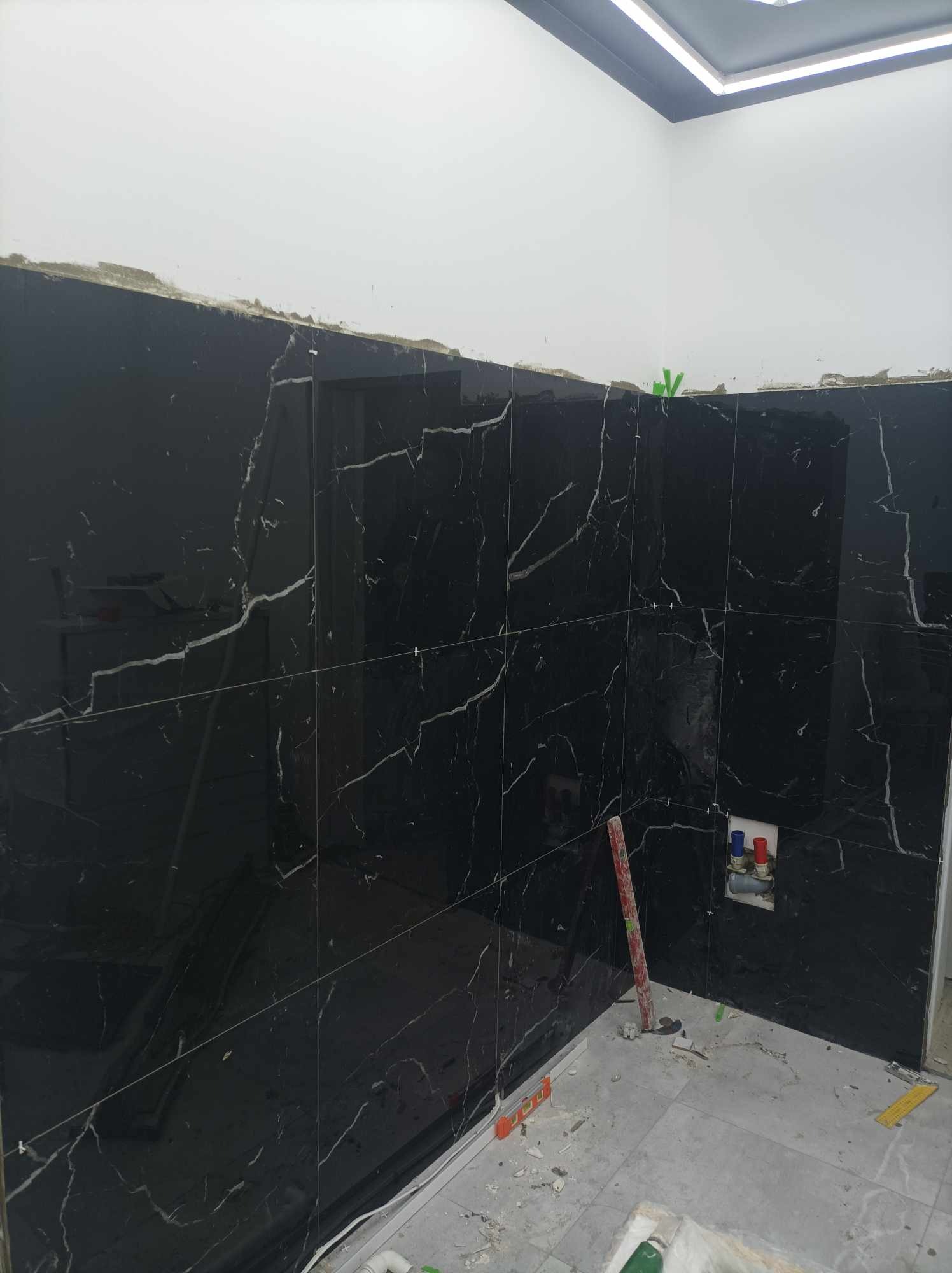 Ściana łazienki w trakcie układania czarnych płytek z białym marmurkowym wzorem, widoczne poziomice i instalacja wodna, ślady zaprawy na płytkach i podłodze.