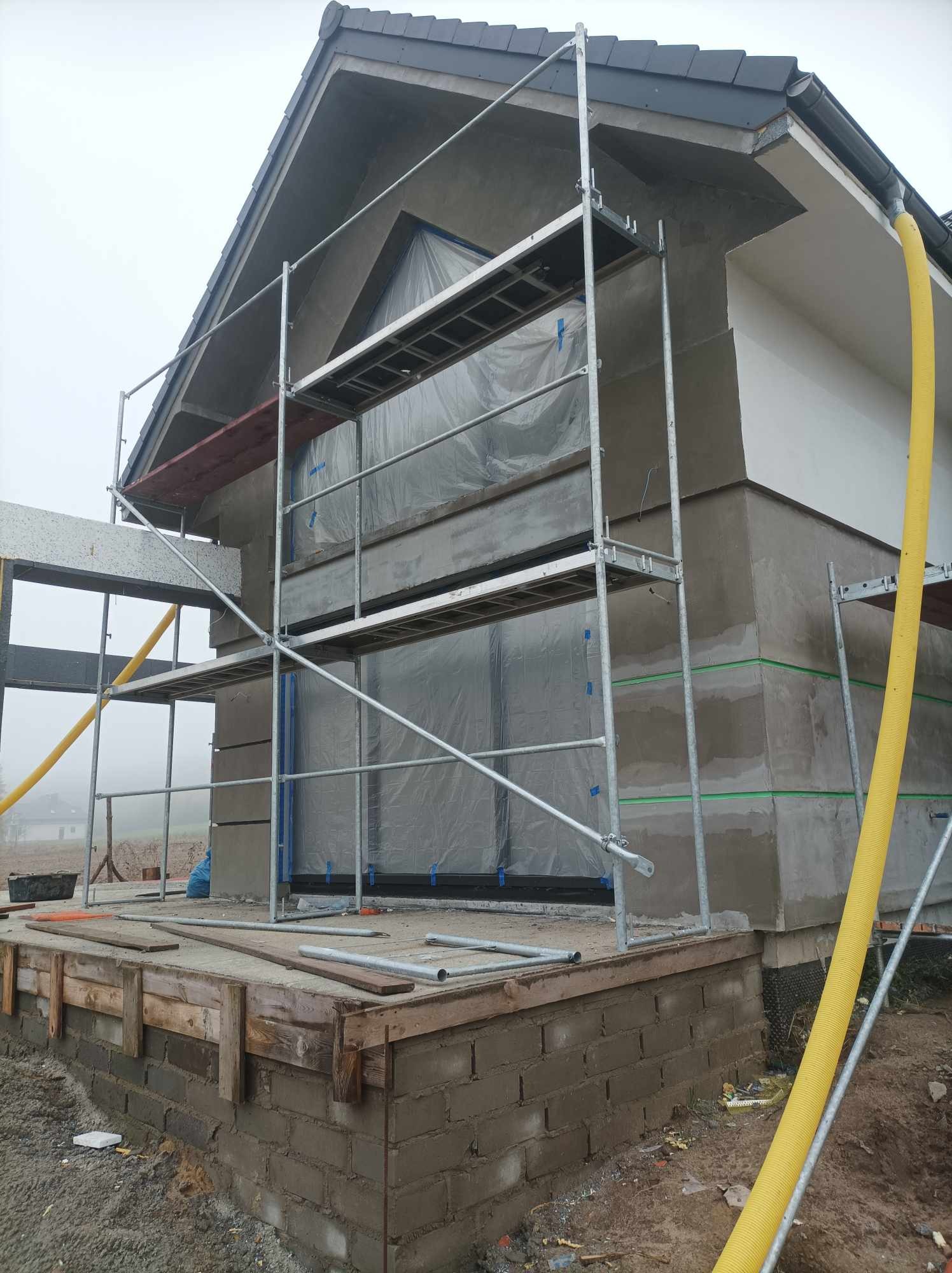 Dom jednorodzinny w trakcie ocieplania, widoczne rusztowania, okna zabezpieczone folią, ściany pokryte szarym tynkiem, żółty przewód wentylacyjny, teren wokół budynku w stanie surowym.