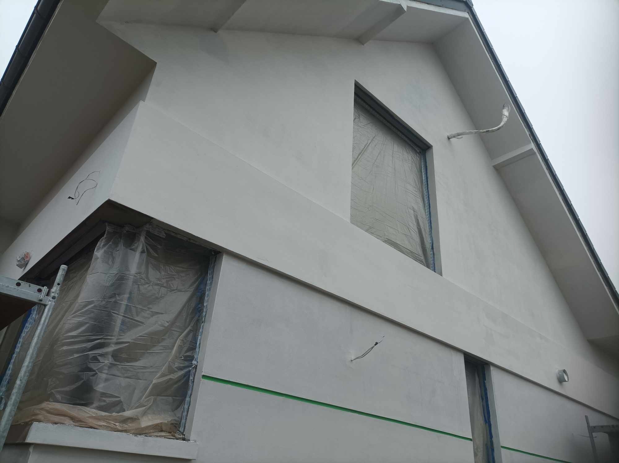 Elewacja domu w trakcie malowania na biało, okna zabezpieczone folią, widoczny fragment rusztowania i wystający kabel.