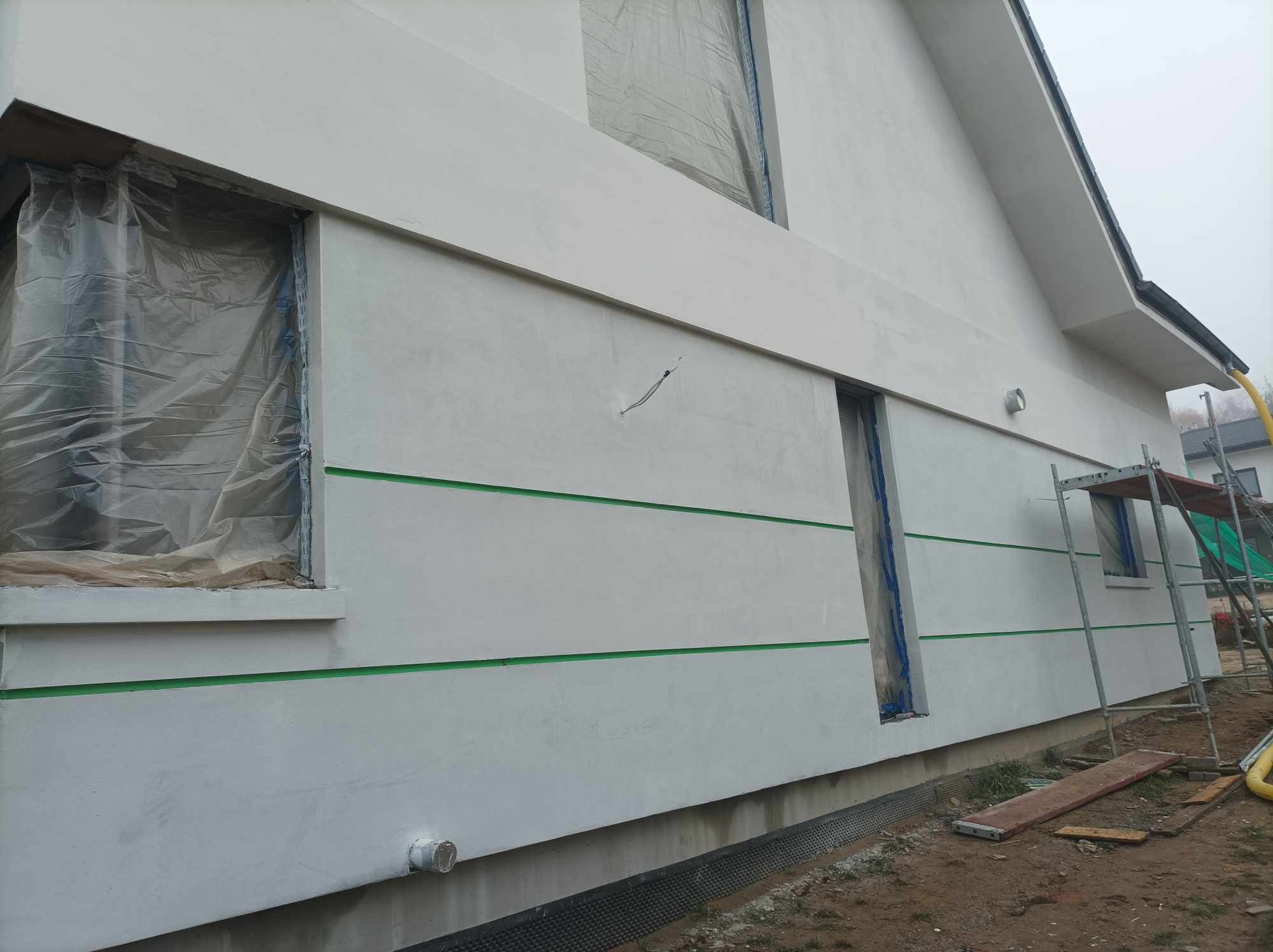 Elewacja domu jednorodzinnego w trakcie malowania, okna zabezpieczone folią, widoczne zielone pasy oddzielające fragmenty elewacji, rusztowanie przy ścianie.