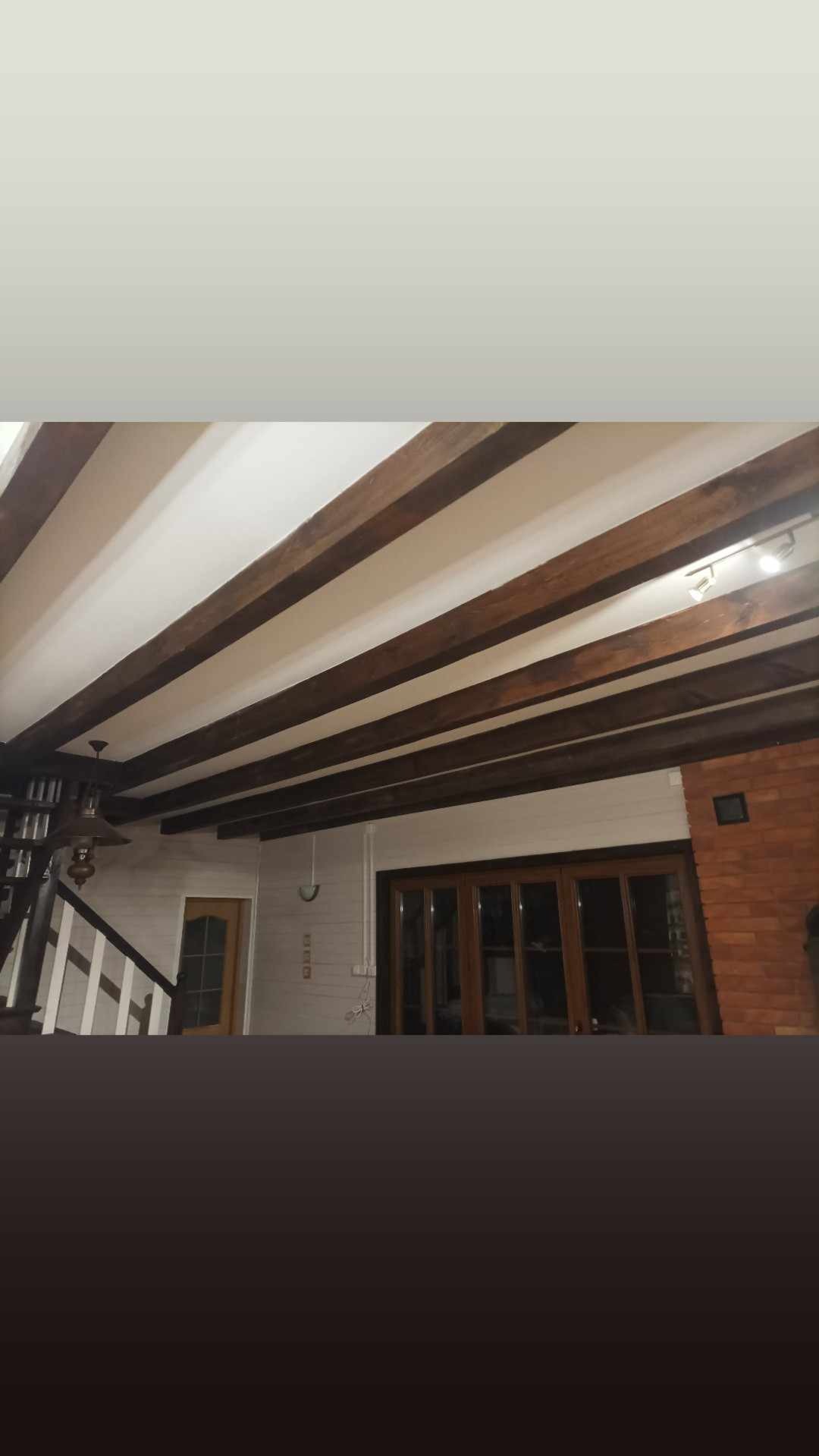 Drewniane belki stropowe w salonie z ceglanym kominkiem, widoczne schody z białymi balustradami i lampa sufitowa.